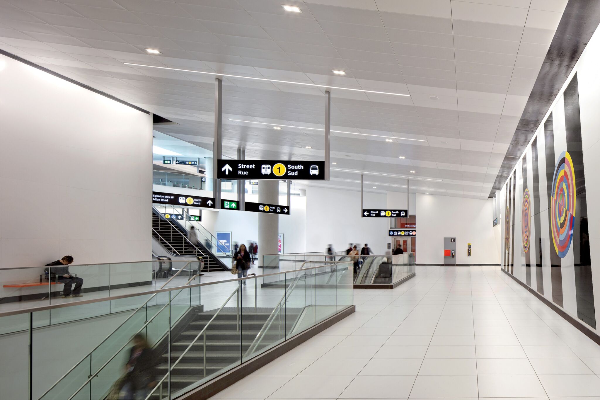 Modern airport interior with white walls, escalators, and signs directing to street, train, and taxi. Several people walking or standing. Large circular artwork decorates the right wall.
