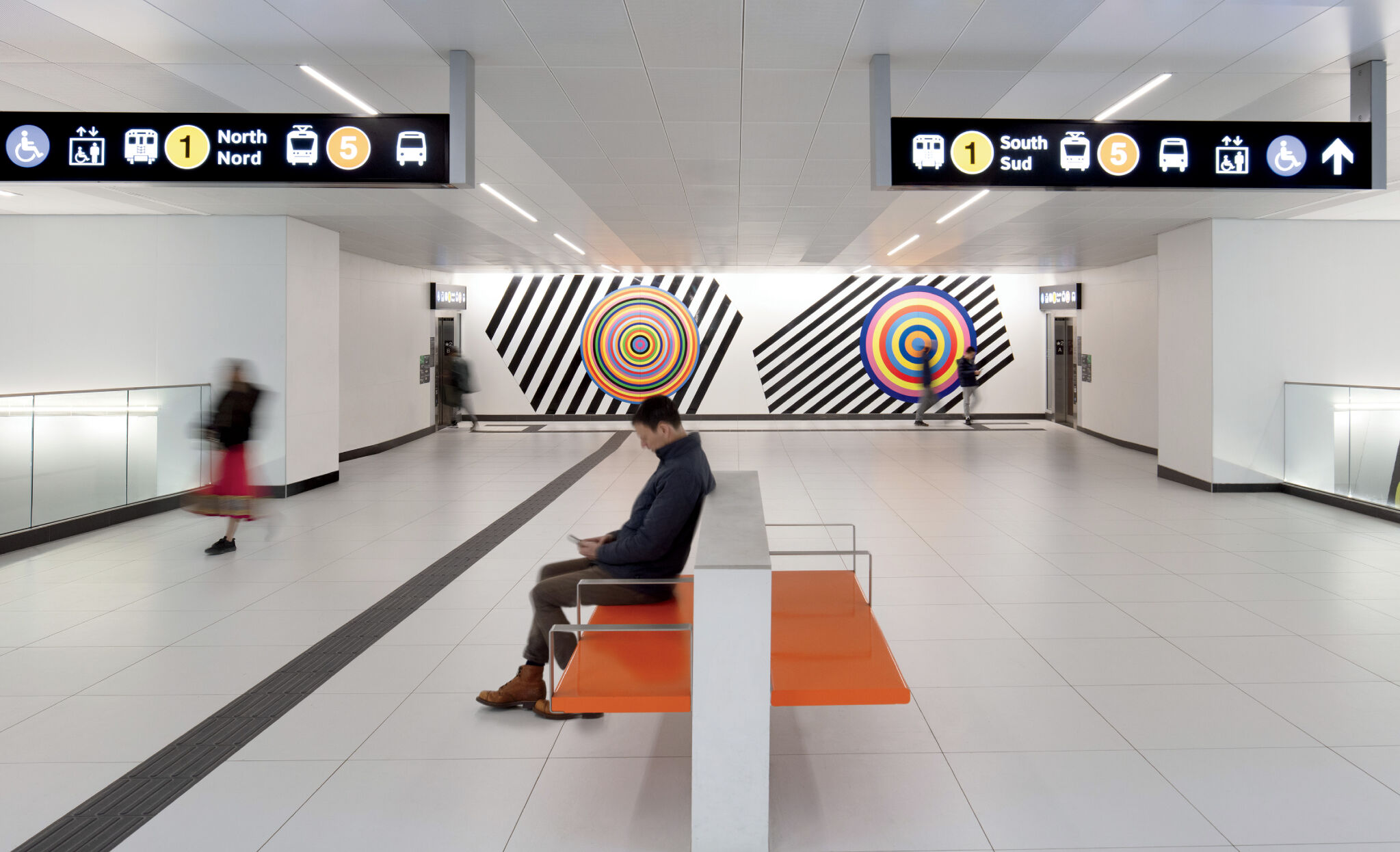 A person sits on an orange bench in a modern subway station, with people walking and colorful geometric art on the walls in the background.