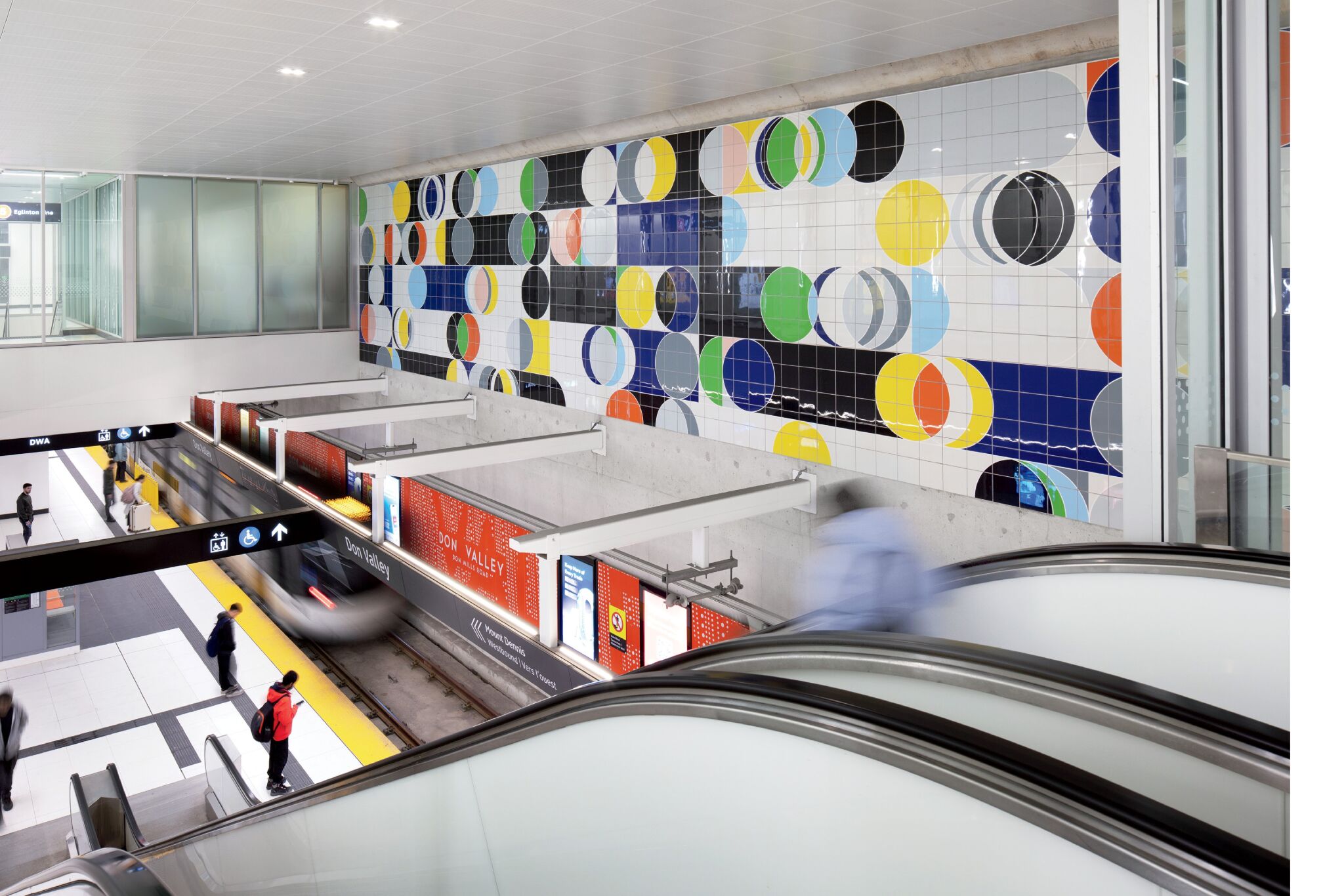 Colorful abstract tile mural decorates the wall above a modern subway platform with escalators, signs, and people waiting or walking.