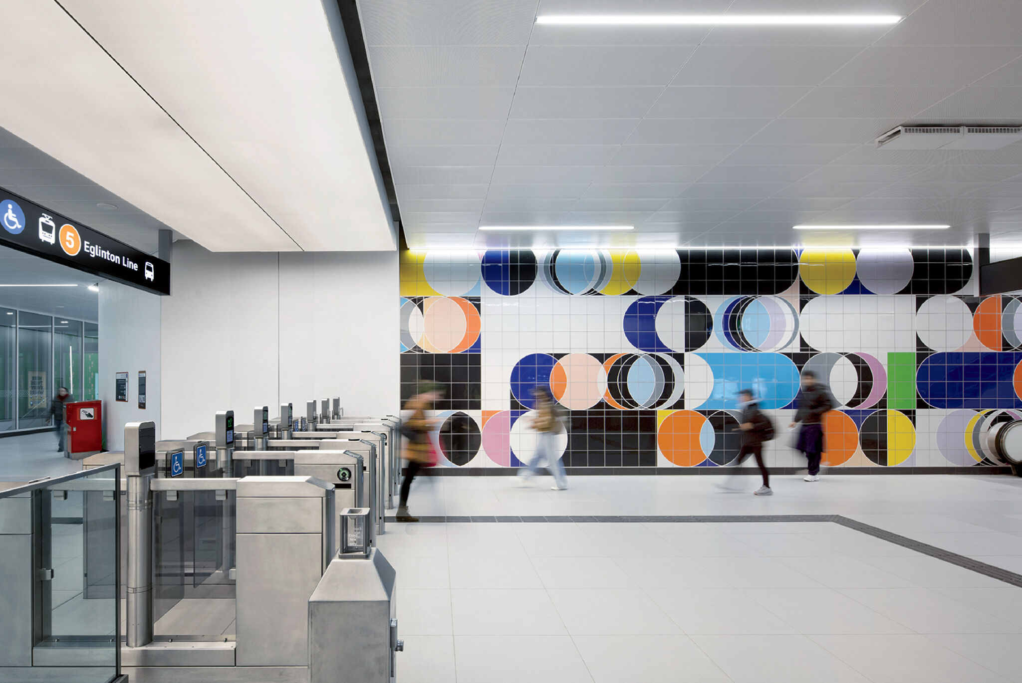 People walk past a colorful geometric mural and fare gates in a modern, brightly lit subway station.
