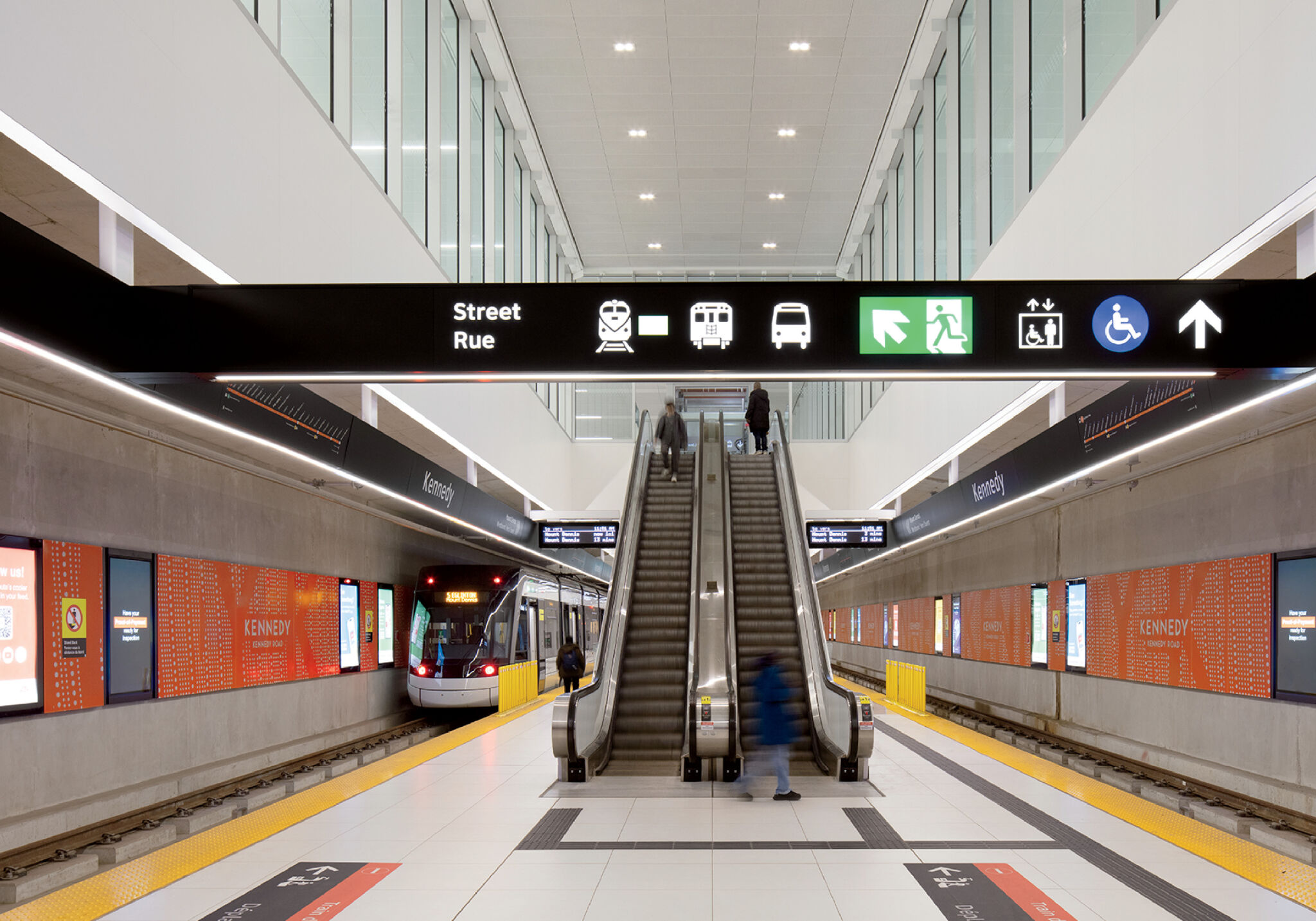 Indoor light rail station with an incoming train on the left, an escalator leading up, and various directional signs overhead.