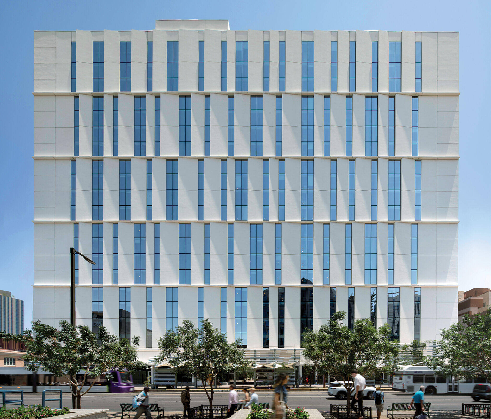A modern white office building with vertical blue glass windows, trees in front, and people sitting on benches along the sidewalk.