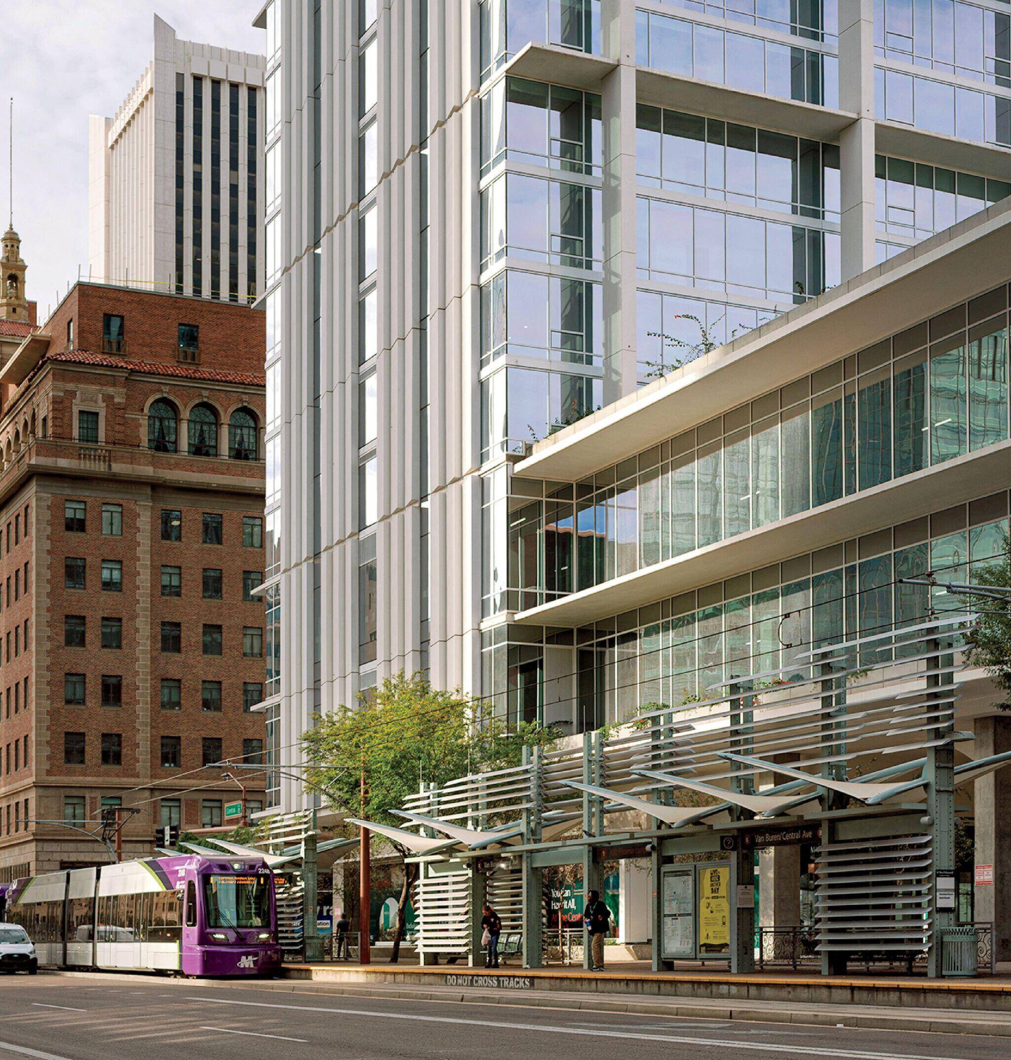 A purple light rail train passes modern glass buildings and older brick architecture on a city street lined with trees.