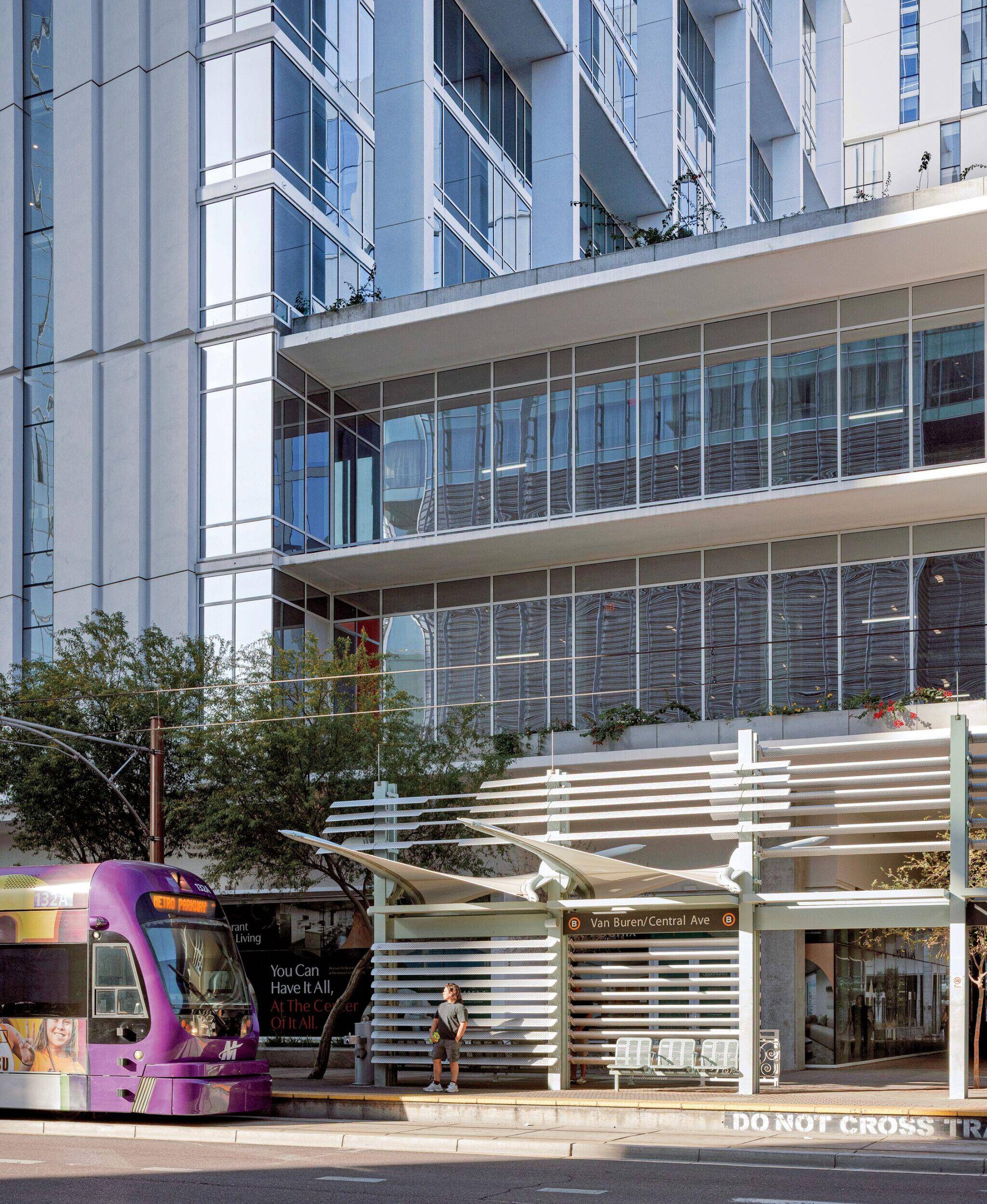 A purple tram stops at Van Buren/Central Ave station in front of a modern glass building; one person stands on the platform.