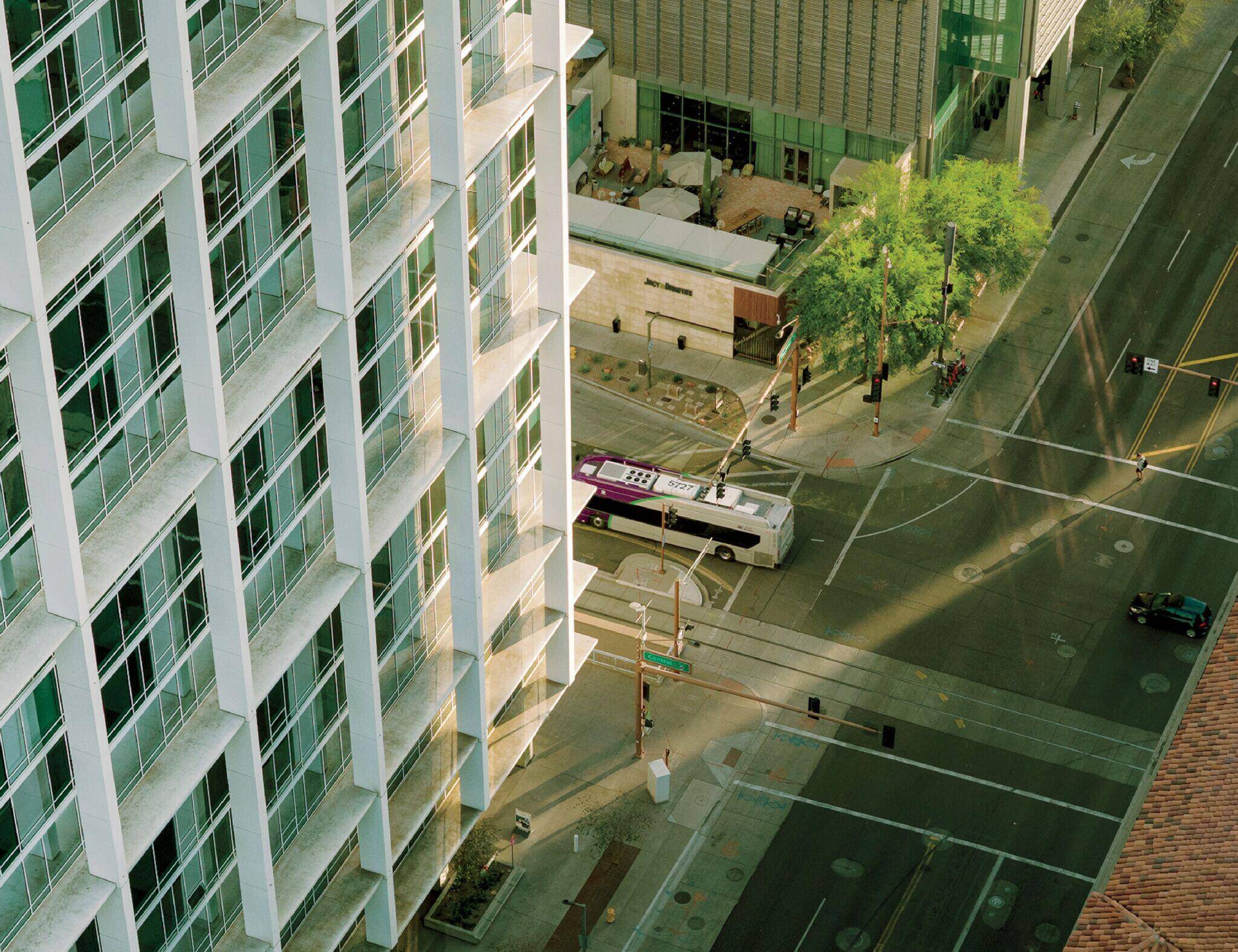 A city street scene viewed from above, showing a white and purple bus stopped near a modern glass building, with some trees and people visible nearby.