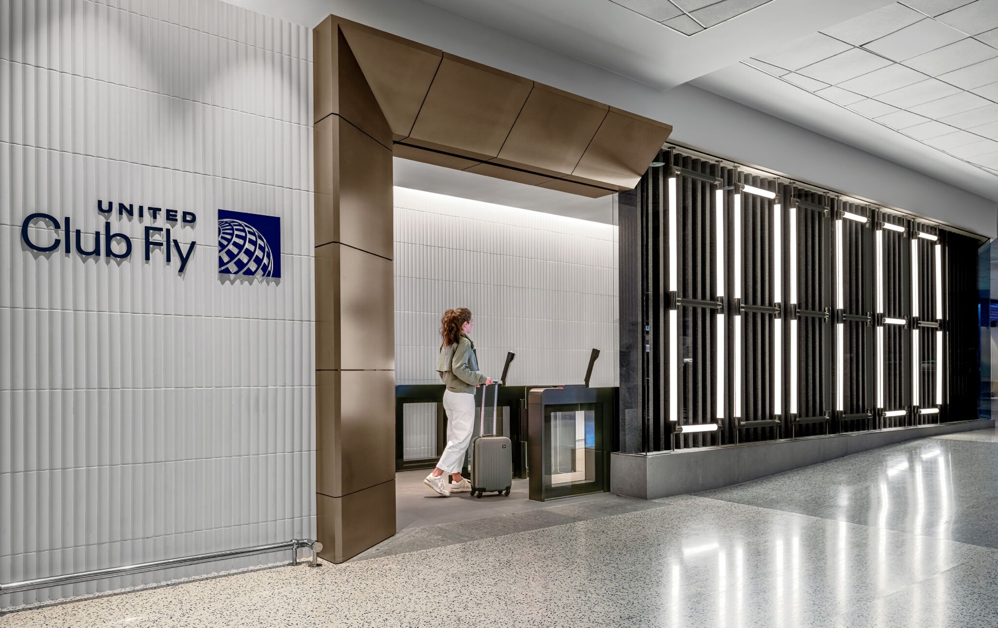 A person with a suitcase stands at the entrance of United Club Fly, an airline lounge, inside an airport terminal.