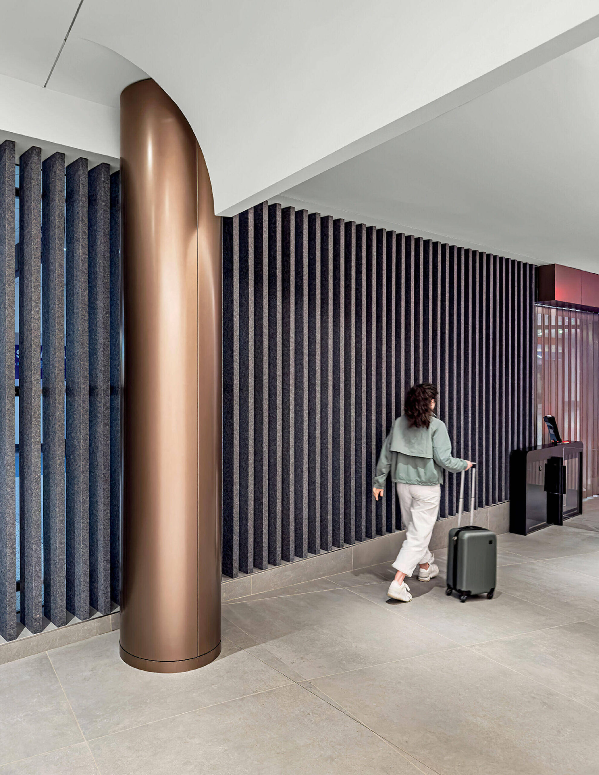 Person with dark hair, light green jacket, and white pants walks indoors pulling a small rolling suitcase past a bronze column and textured dark vertical wall panels.
