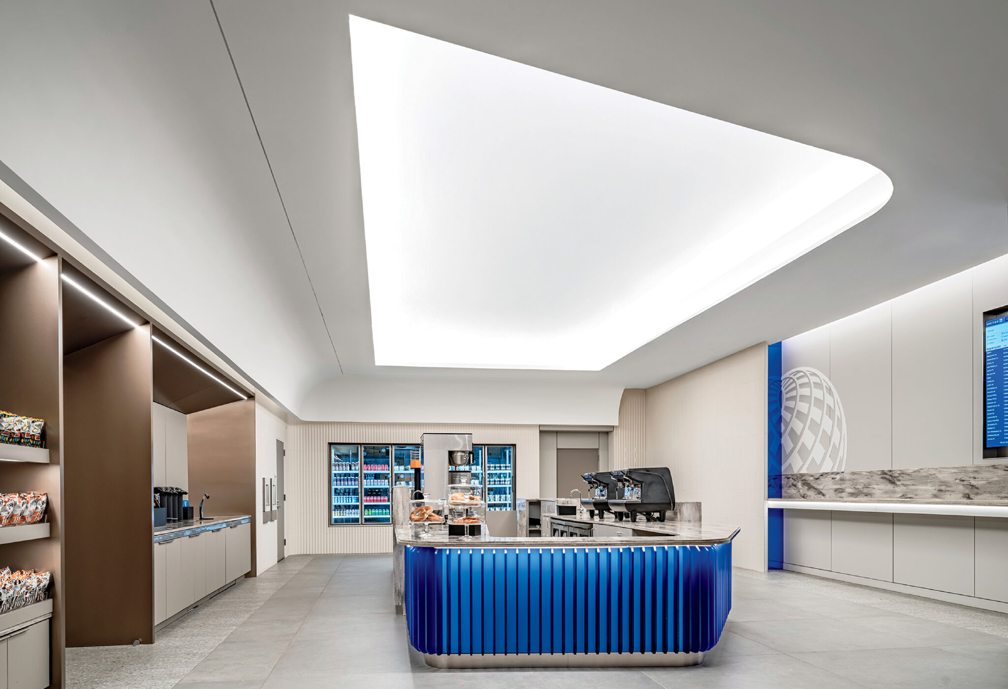 Modern airport lounge with a blue-accented snack and coffee counter, shelves with drinks and snacks, and a digital flight information display on the wall.
