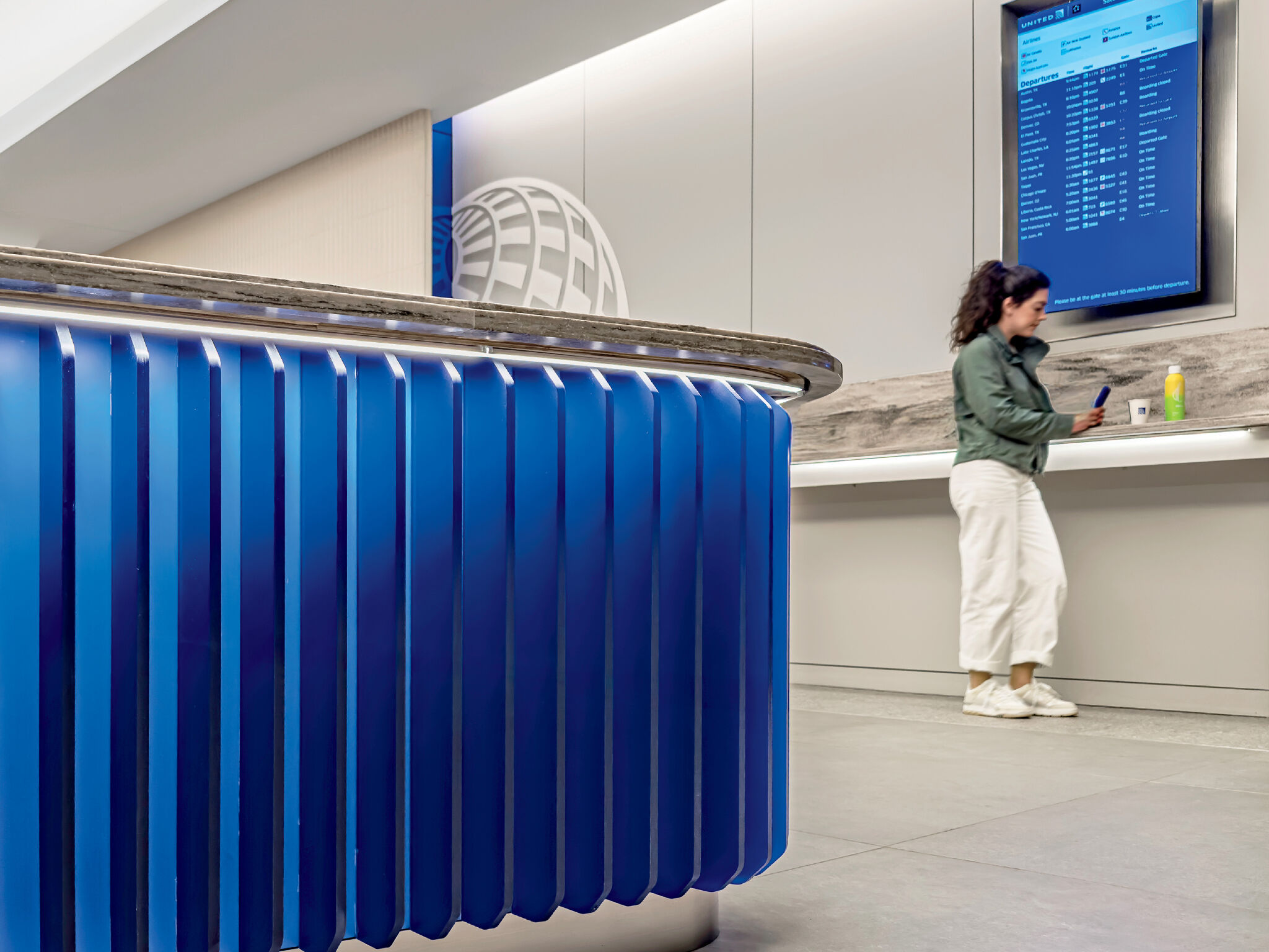 A woman stands at a counter in an airport terminal, looking at her phone. A digital flight information board is mounted on the wall nearby.