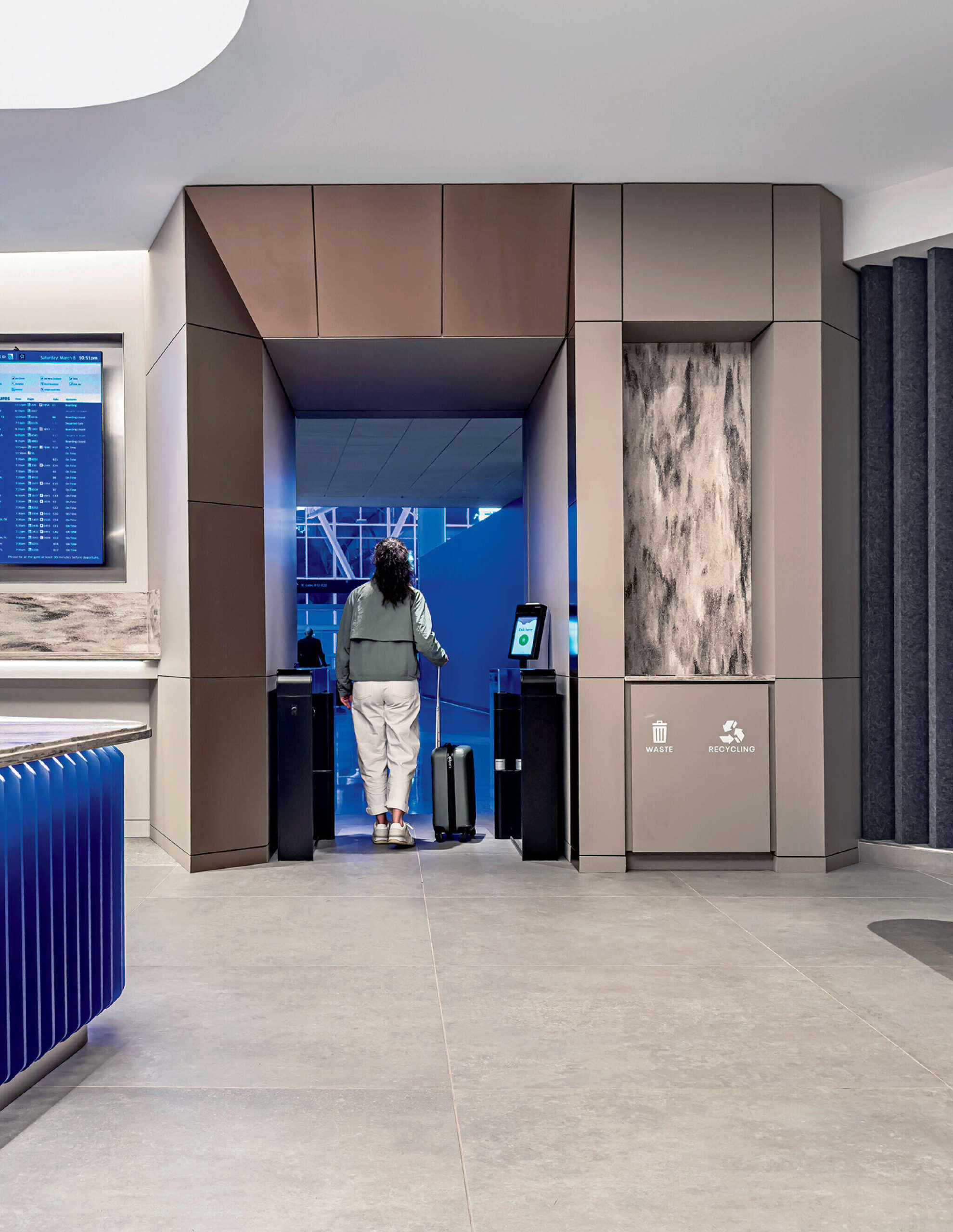 Person with a suitcase walking through an airport boarding gate, facing away from the camera, with digital screens and recycling bins visible.