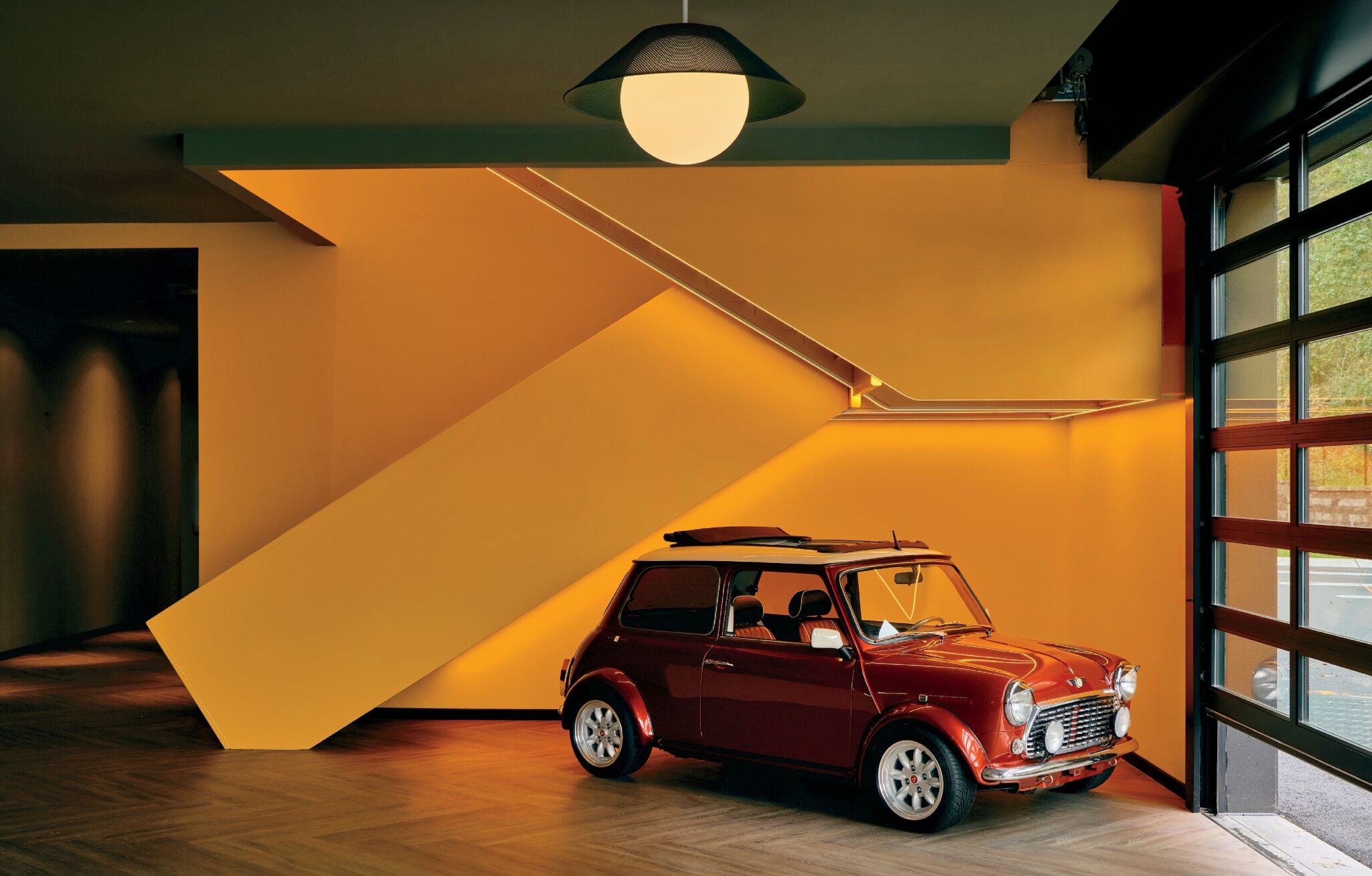 A classic red Mini Cooper is parked indoors beside a yellow staircase with modern lighting and a large window.