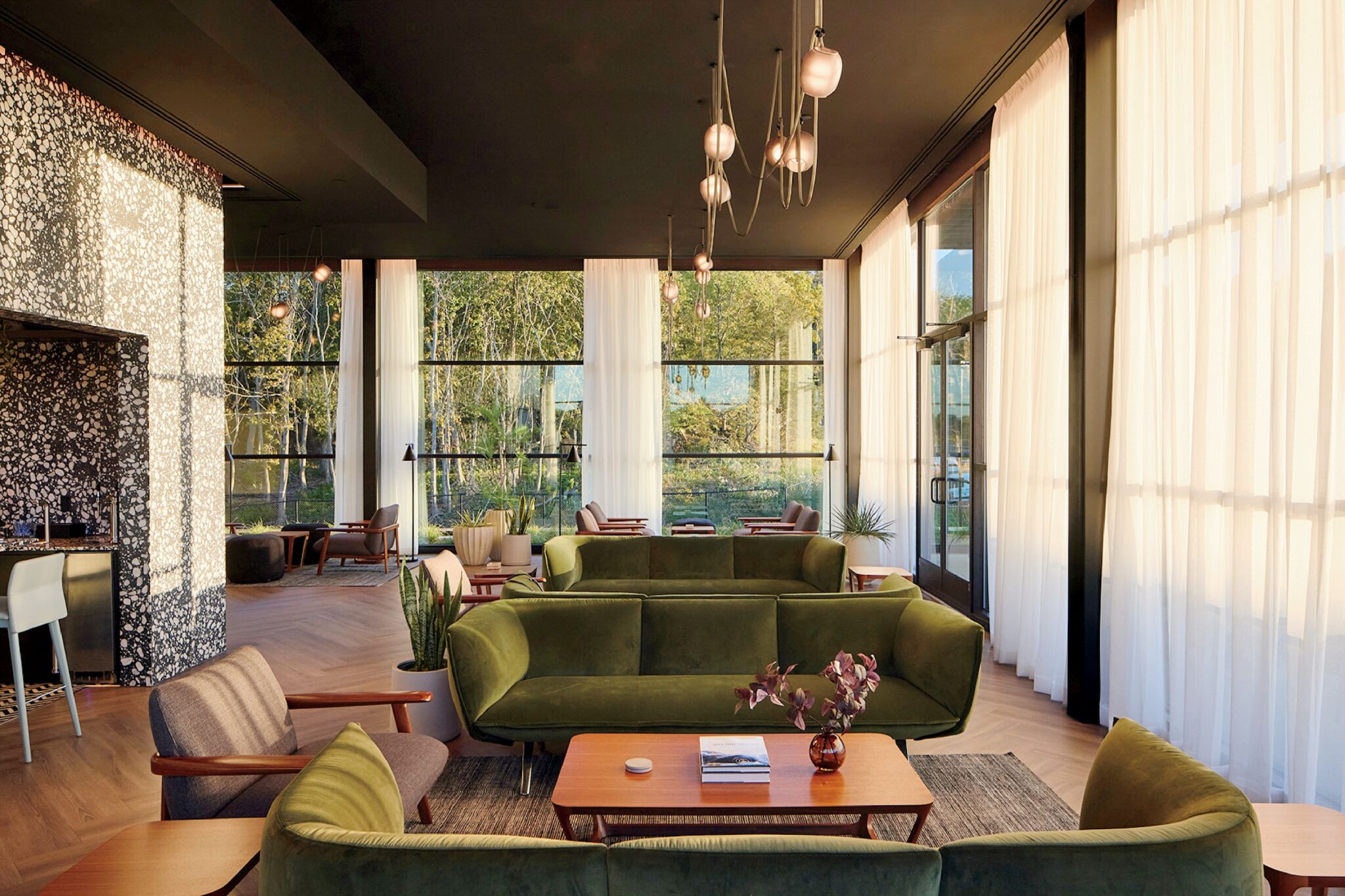Modern lounge with green sofas, wooden tables, armchairs, large windows with sheer curtains, and pendant lights, overlooking a garden area outside.