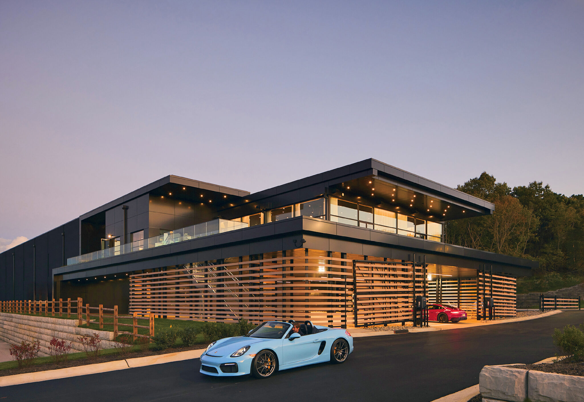 Modern two-story building with large glass windows and horizontal wooden slats. A light blue convertible and a red car are parked outside on a paved driveway at sunset.