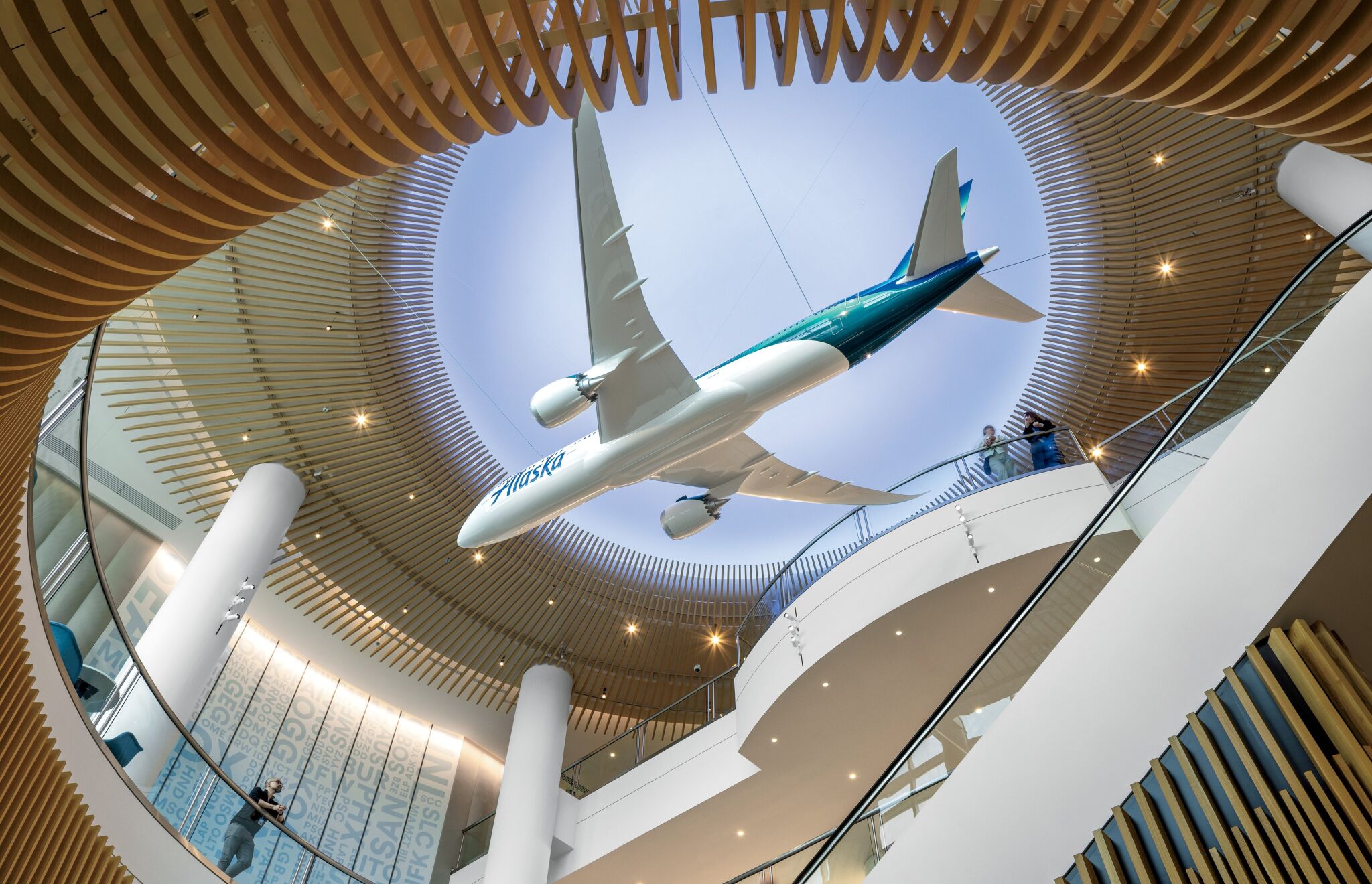 View from below of a suspended airplane model in a modern, multi-level building with curved balconies and a circular skylight.