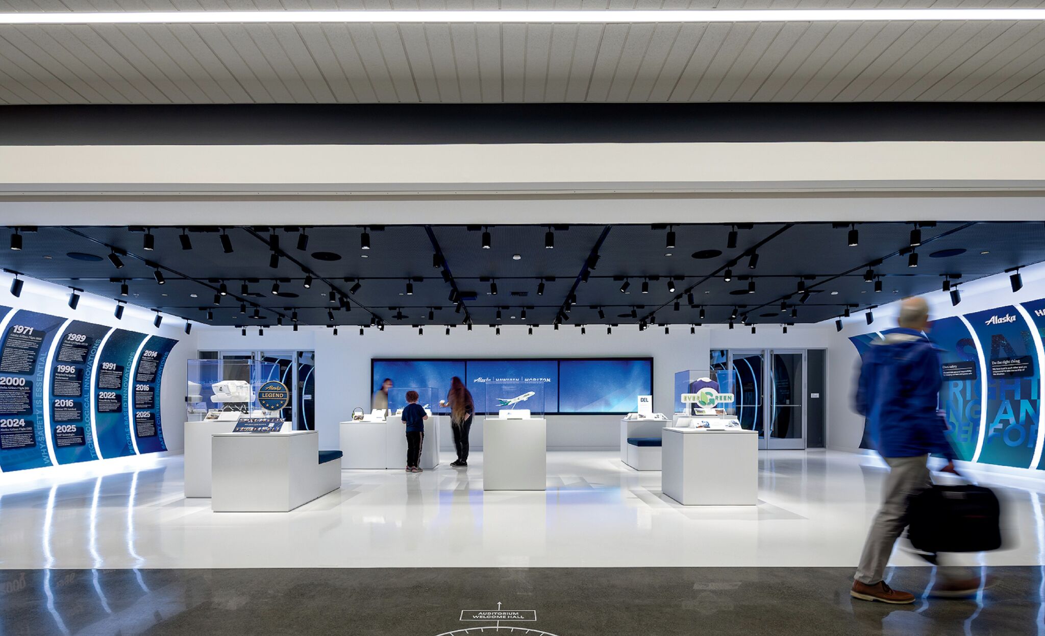 A modern exhibition space features interactive displays, informational panels, and visitors observing models and exhibits under bright lighting.
