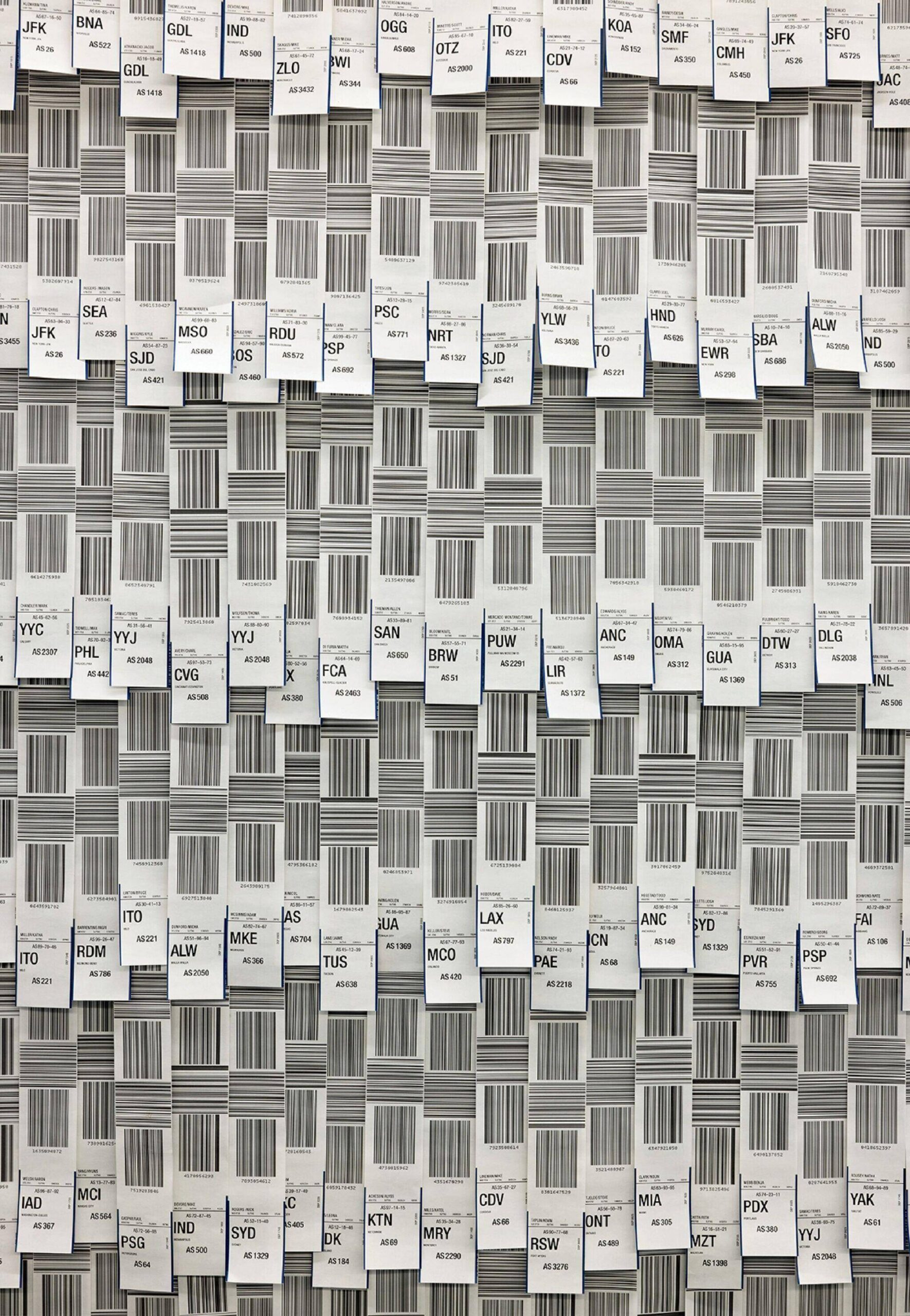 Numerous airline baggage tags with barcodes and three-letter airport codes are arranged vertically and horizontally in a dense grid pattern on a wall.