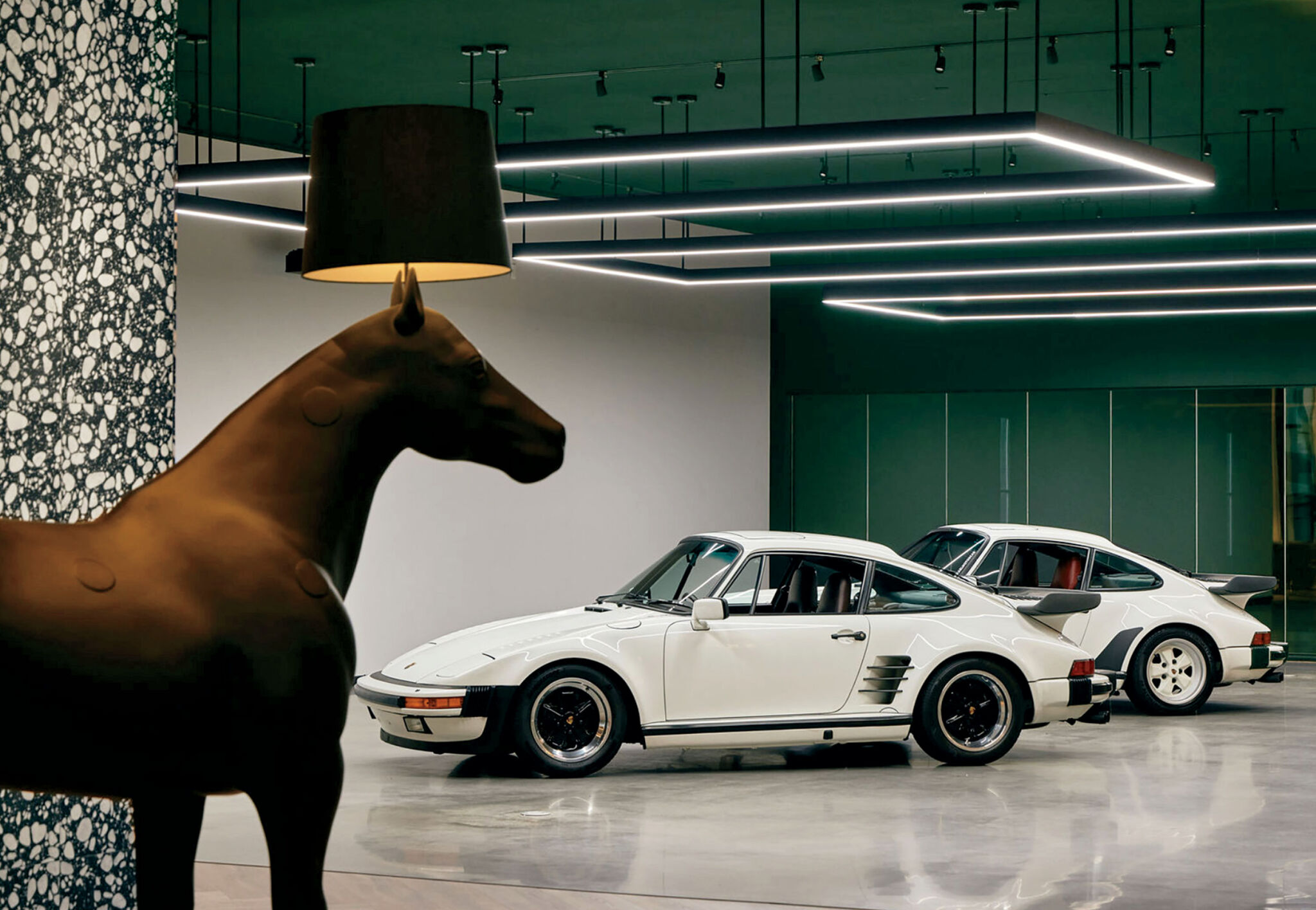 Two white vintage sports cars are parked indoors on a polished concrete floor, with a large horse-shaped lamp in the foreground and modern ceiling lights above.