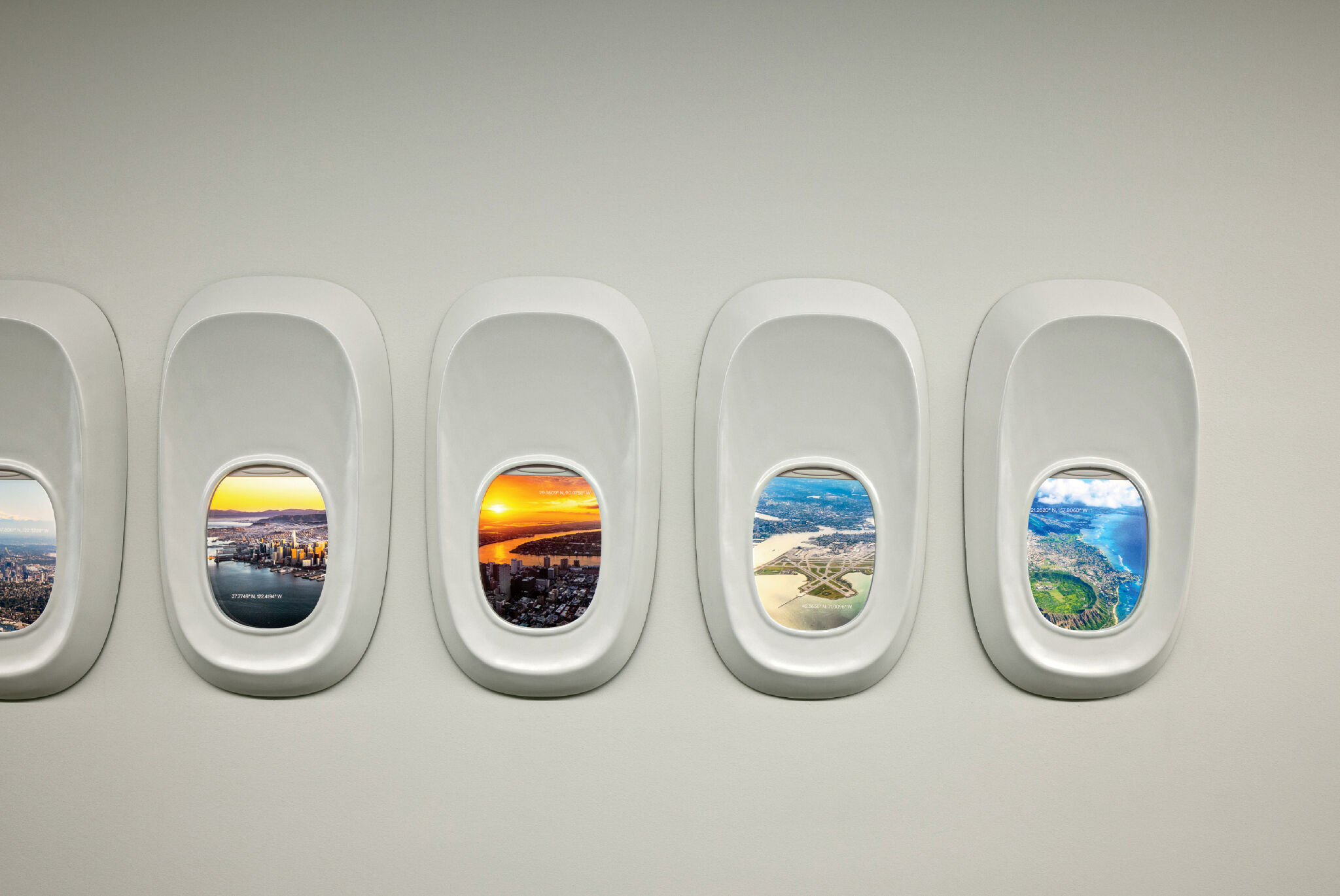 Four airplane windows on a plain wall, each showing a different aerial view of landscapes and cityscapes through the glass.