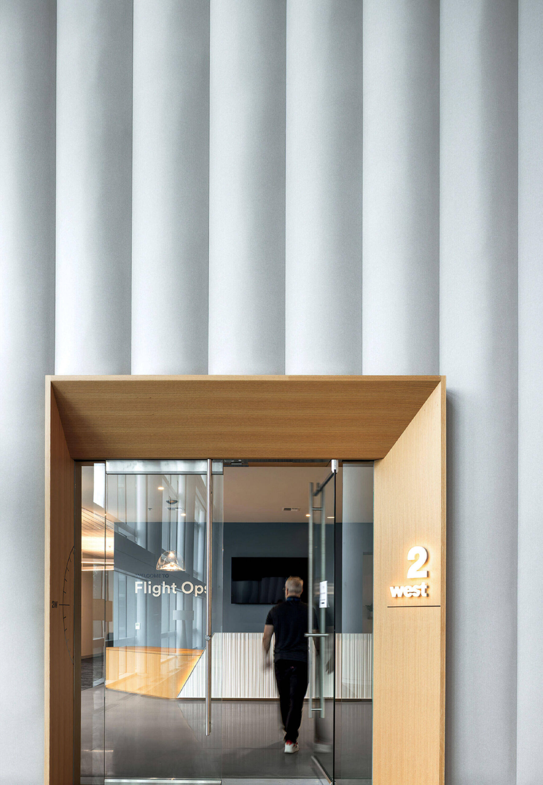 Person walking through a glass door labeled “Flight Op,” under a light wood frame, with a “2 West” sign and tall, vertical wall panels in the background.