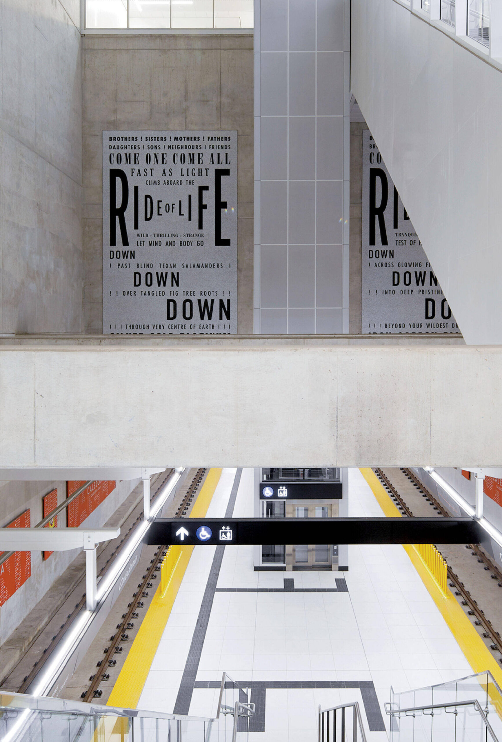 Modern subway station with empty train tracks, concrete walls, and large graphic text posters on the upper wall. Overhead signs for elevator and accessibility are visible.