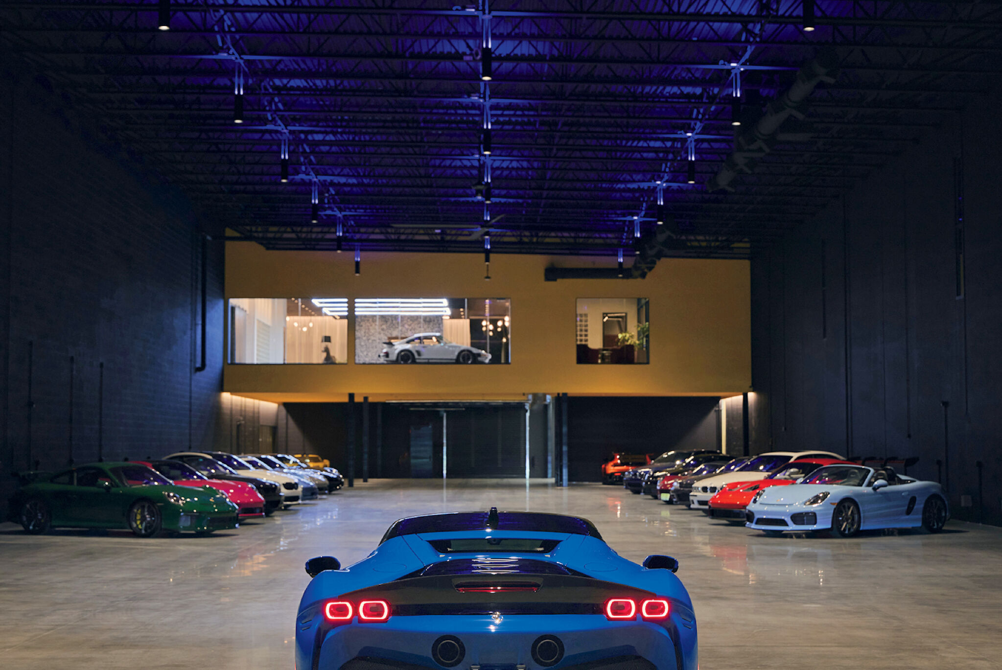 A blue sports car is parked inside a spacious garage, with various other colorful sports cars lined up along the walls and a glass-walled room visible above.
