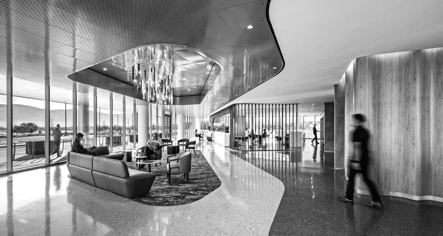 Modern office lobby with seating area, large windows, and contemporary lighting—an environment reminiscent of the sophisticated headquarters often favored by healthcare giants. Several people are sitting, while one person walks across the polished floor. Black and white image.