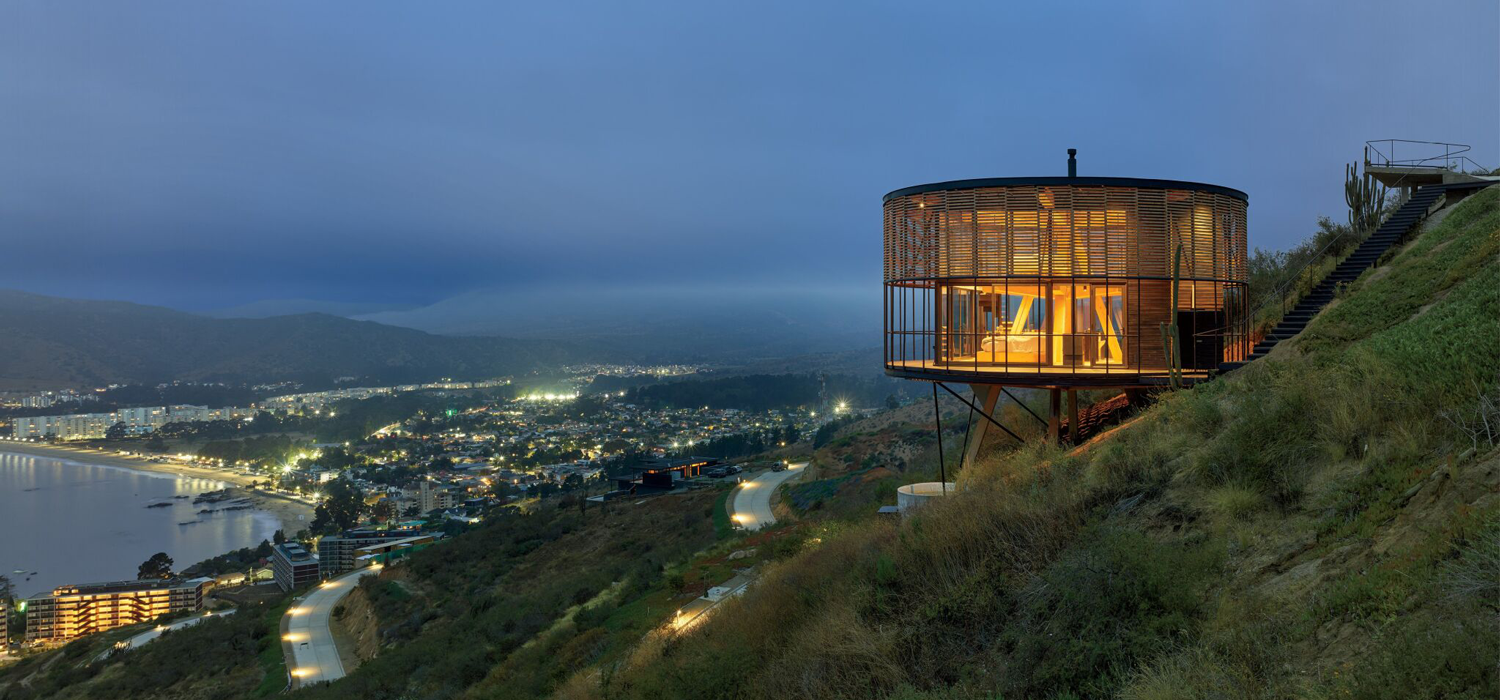 Round, modern house with floor-to-ceiling windows overlooks a coastal city at dusk, situated on a hillside with illuminated roads below.