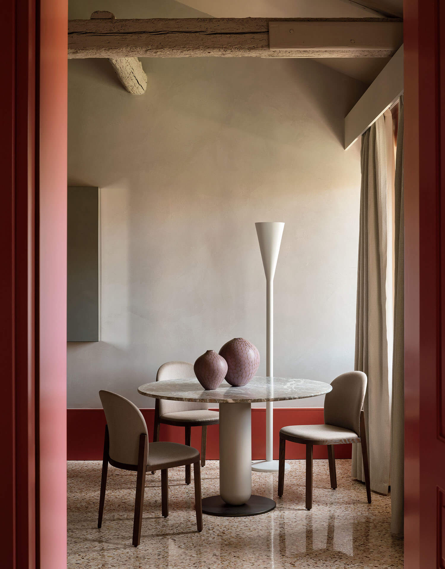 A round marble table with three chairs, two ceramic vases, a tall white floor lamp, and neutral walls, seen through a red-framed doorway.