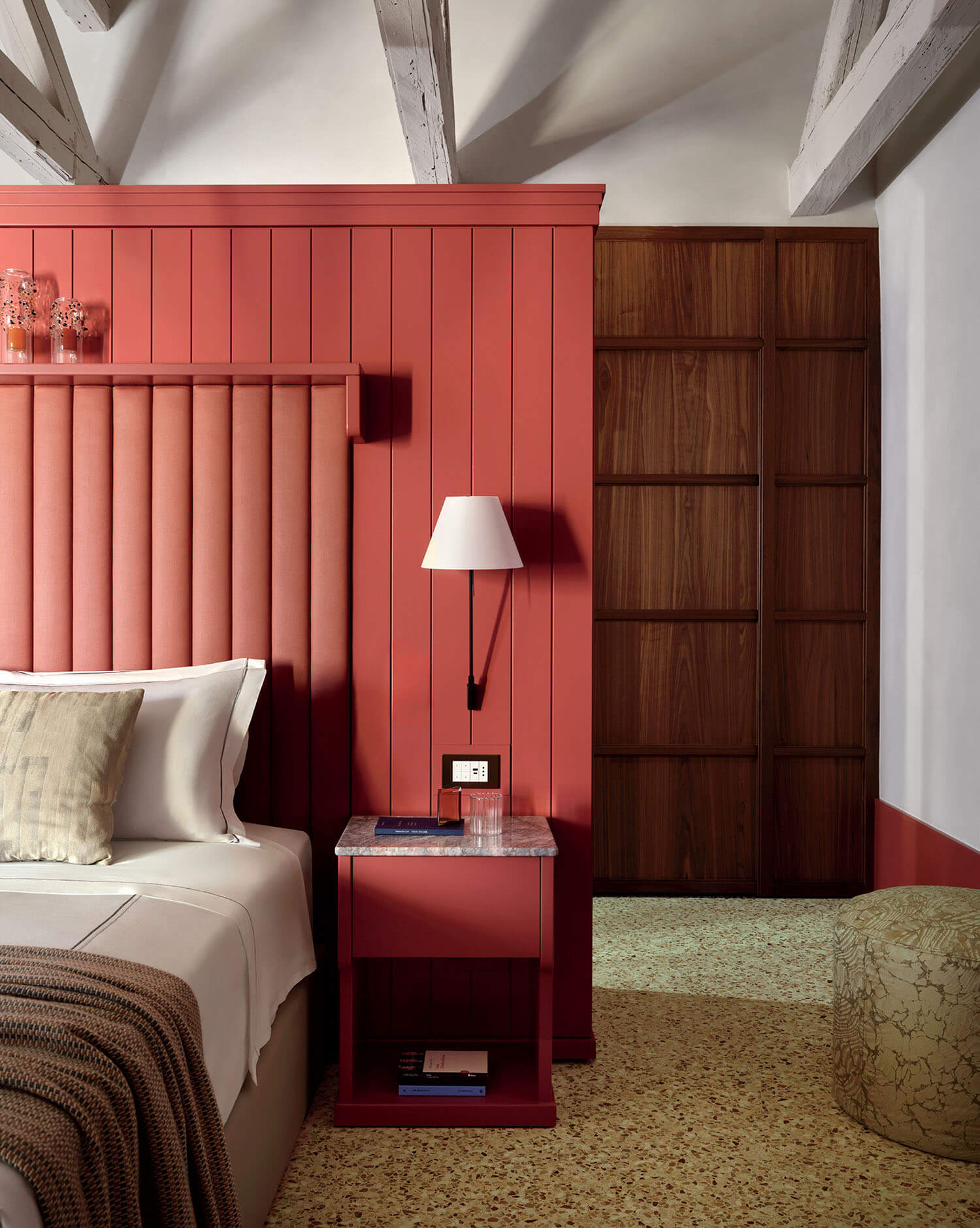 Bedroom with a red paneled accent wall, upholstered headboard, bedside table with lamp, neutral bedding, wooden door, and exposed ceiling beams.