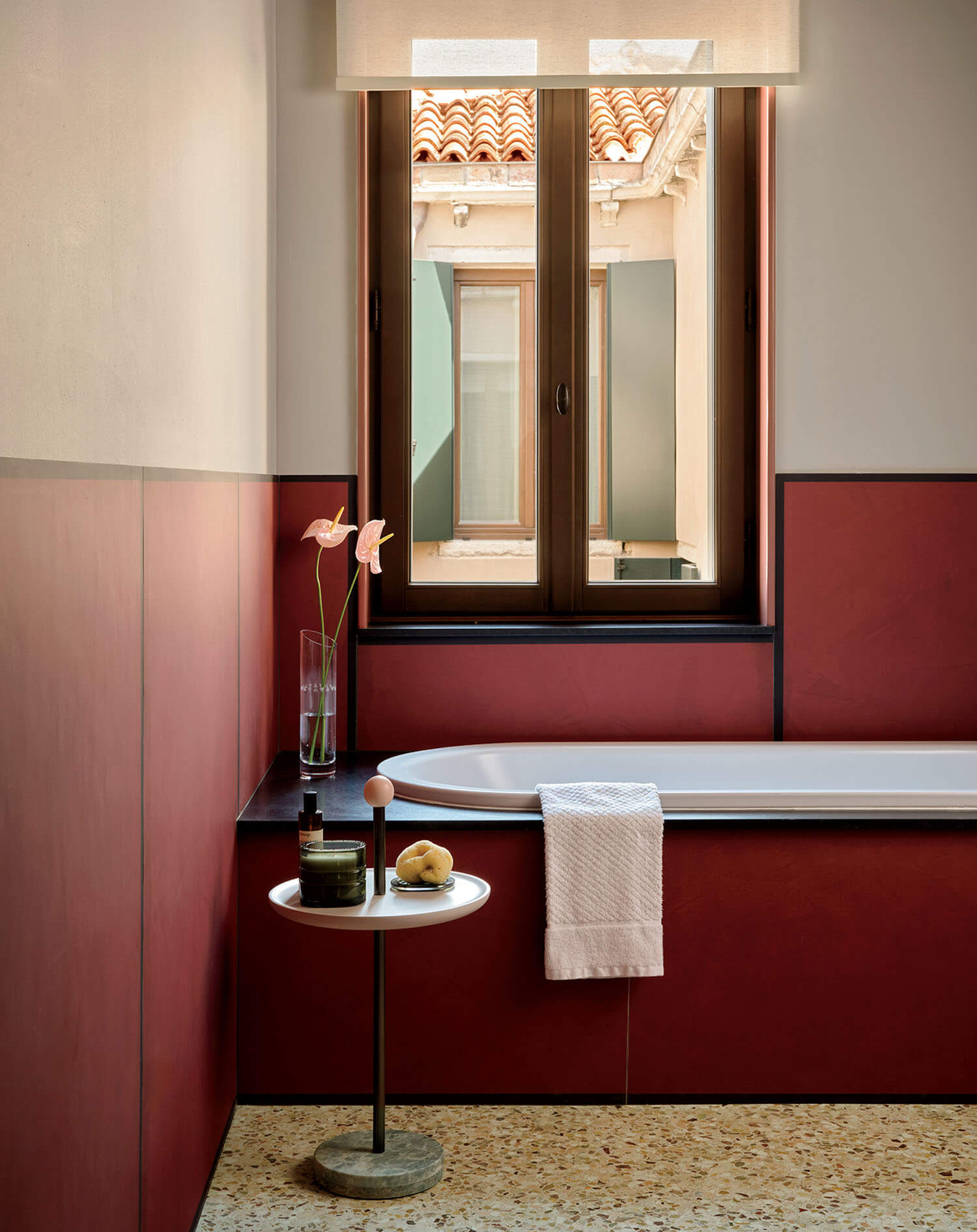 Red-walled bathroom with a white tub, towel, small round side table holding toiletries and flowers, and a window showing tiled rooftops outside.