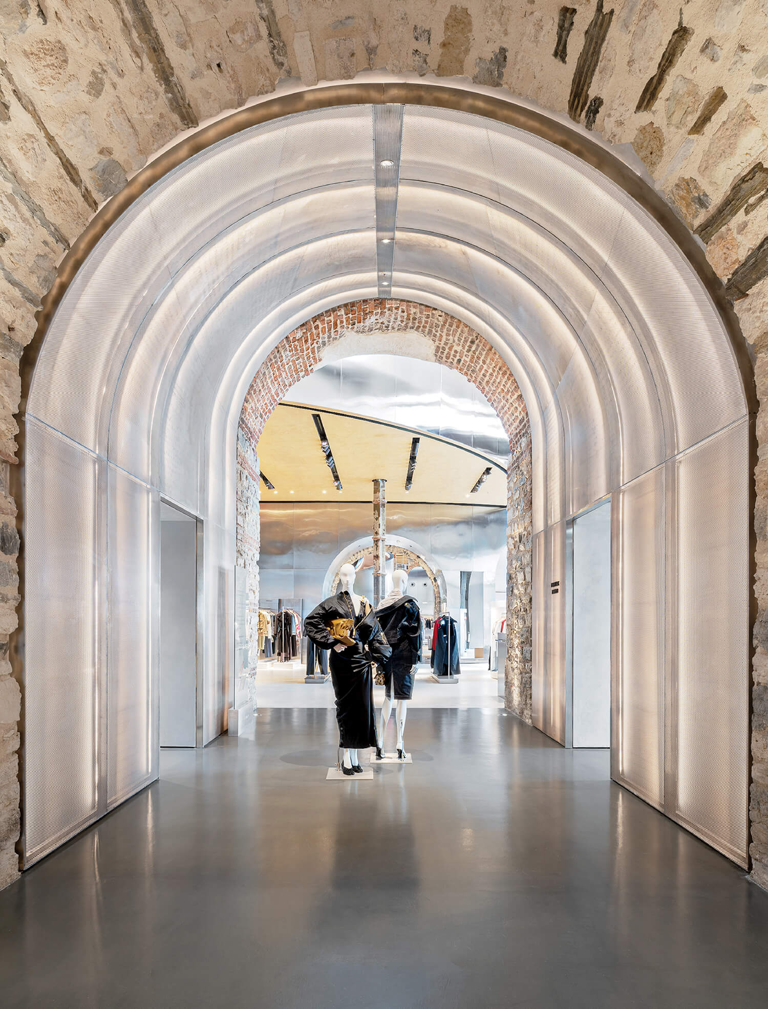 A modern clothing store interior with arched stone walls, a polished floor, and two mannequins dressed in black and white outfits at the center.