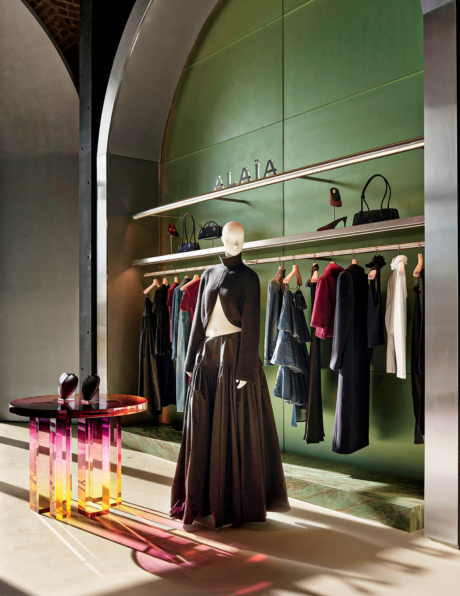 Mannequin in black outfit stands by a round table and display shelves with clothing, bags, and shoes in a boutique with green walls and the sign “Alaïa.”.