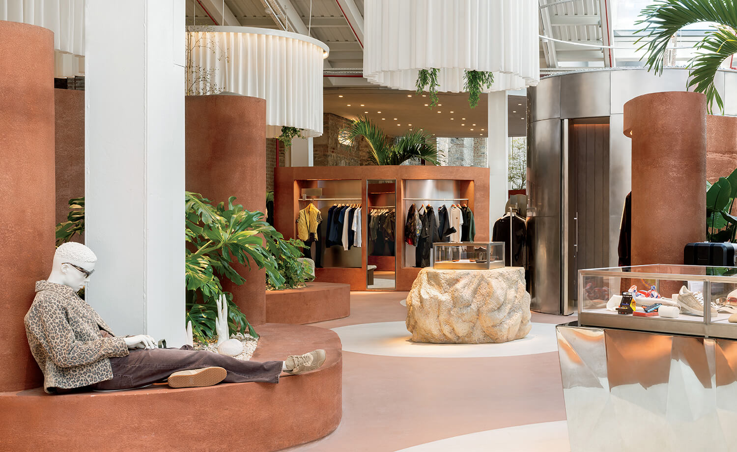Modern clothing store interior with curved brown walls, clothing racks, glass display cases, plants, and a seated mannequin wearing a jacket and hat.