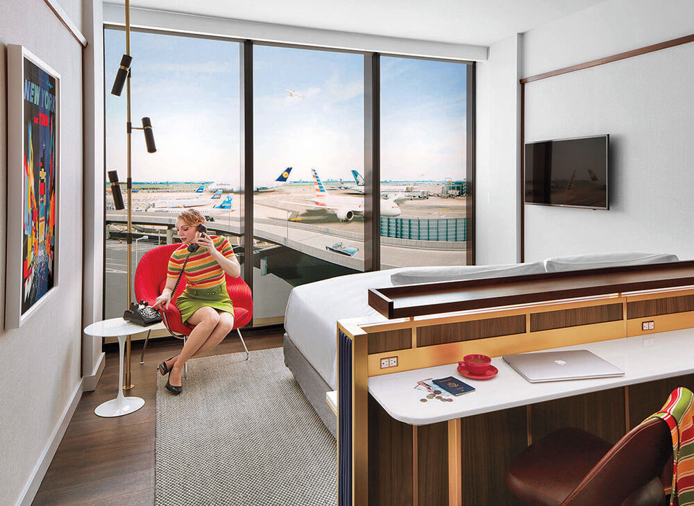 A woman sits in a hotel room with floor-to-ceiling windows overlooking an airport runway, with planes visible outside.