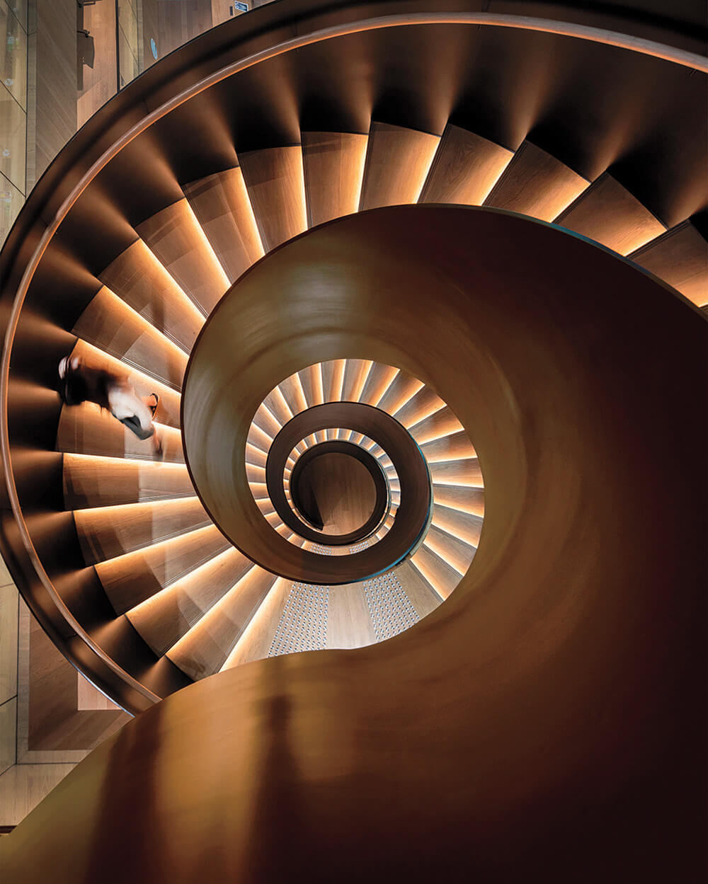 A spiral staircase with illuminated steps viewed from above, with a person walking down. The lighting highlights the curves and structure of the staircase.