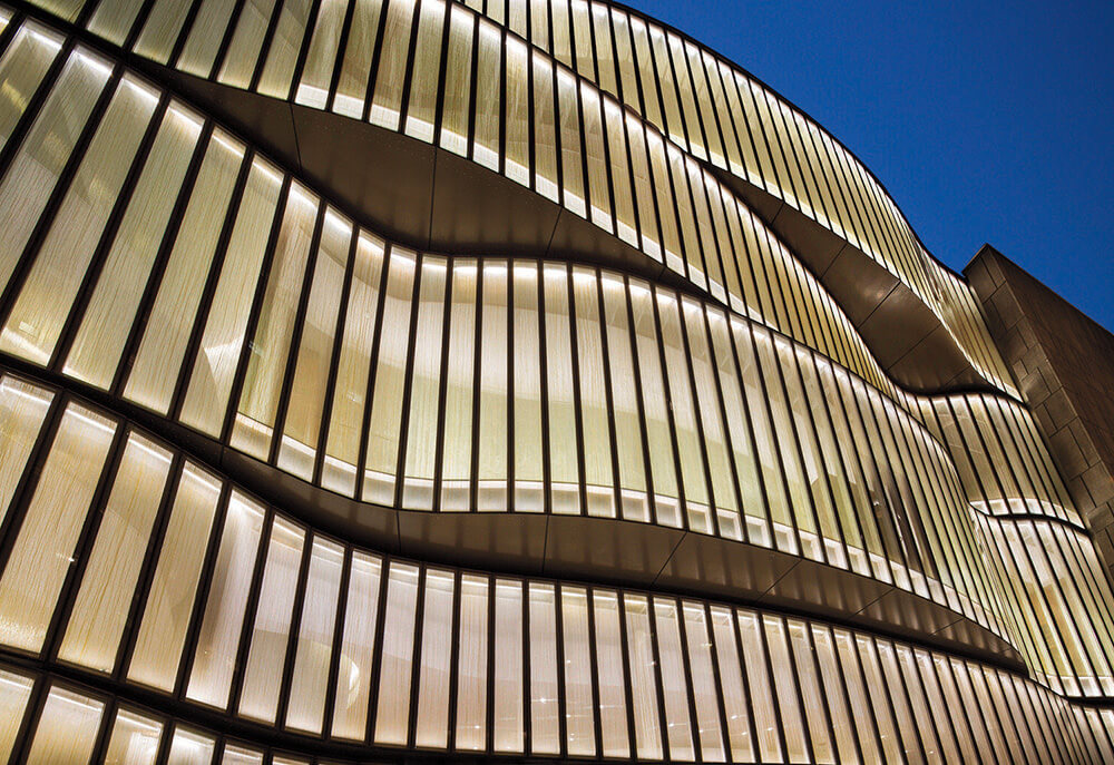 Modern building with wavy, illuminated glass facade viewed from below against a clear evening sky.