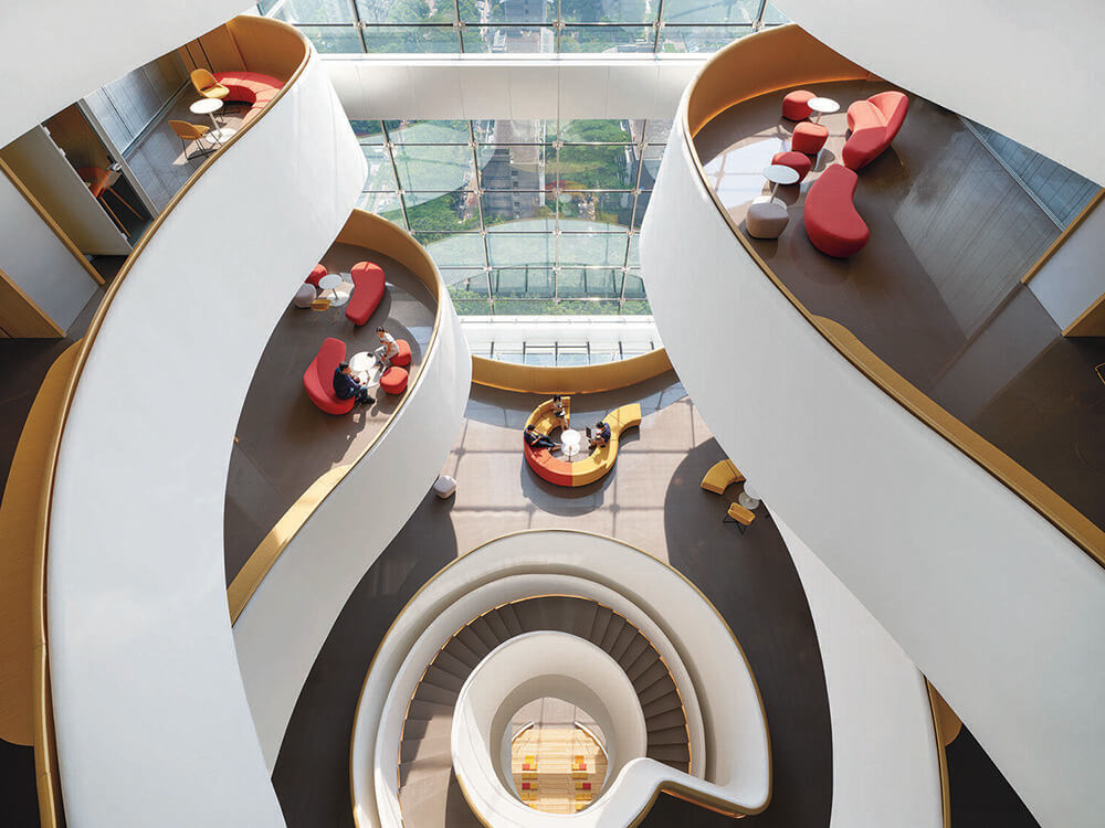 A multi-story atrium with curved walkways, red seating areas, and a central spiral staircase, viewed from above. Large windows provide natural light.