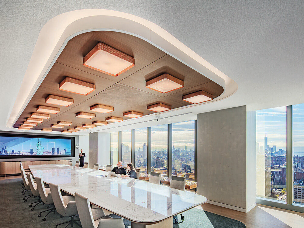 Modern conference room with a long marble table, multiple chairs, unique ceiling lights, and large windows showing a city skyline. Two people sit at the table; one person stands near a large screen.