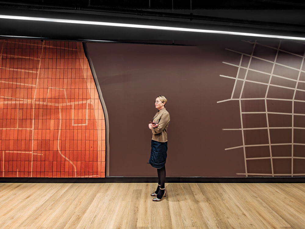 A woman stands on a wooden floor in front of a large wall with abstract map-like designs in orange and brown tones.