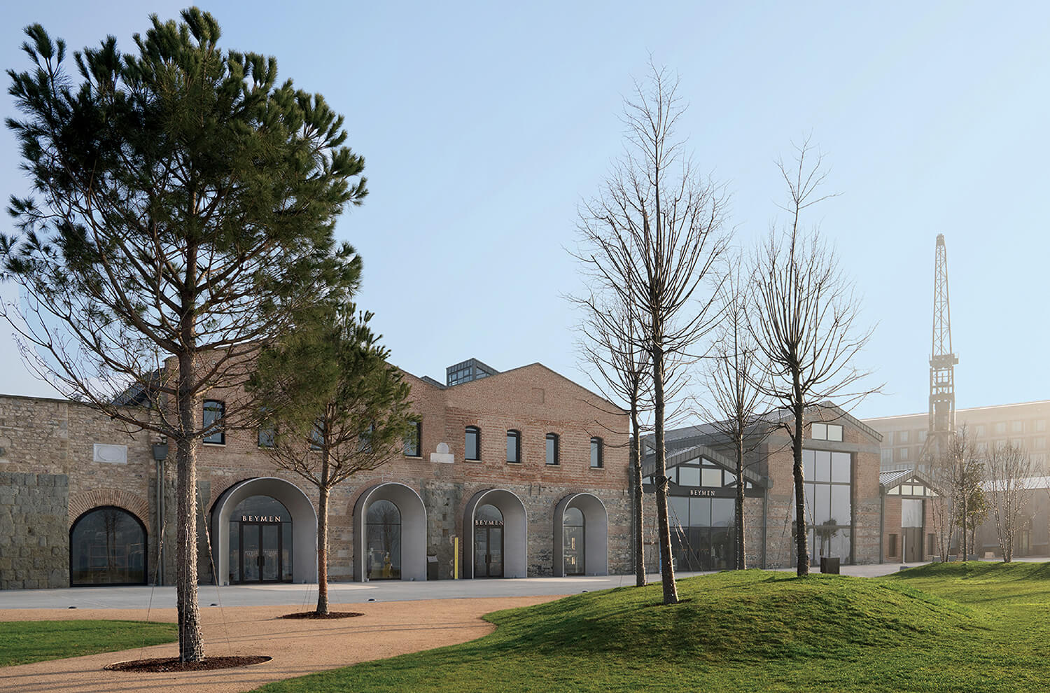 Exterior of a renovated industrial building with arched windows, trees on a grassy lawn, and a crane visible in the background under a clear sky.