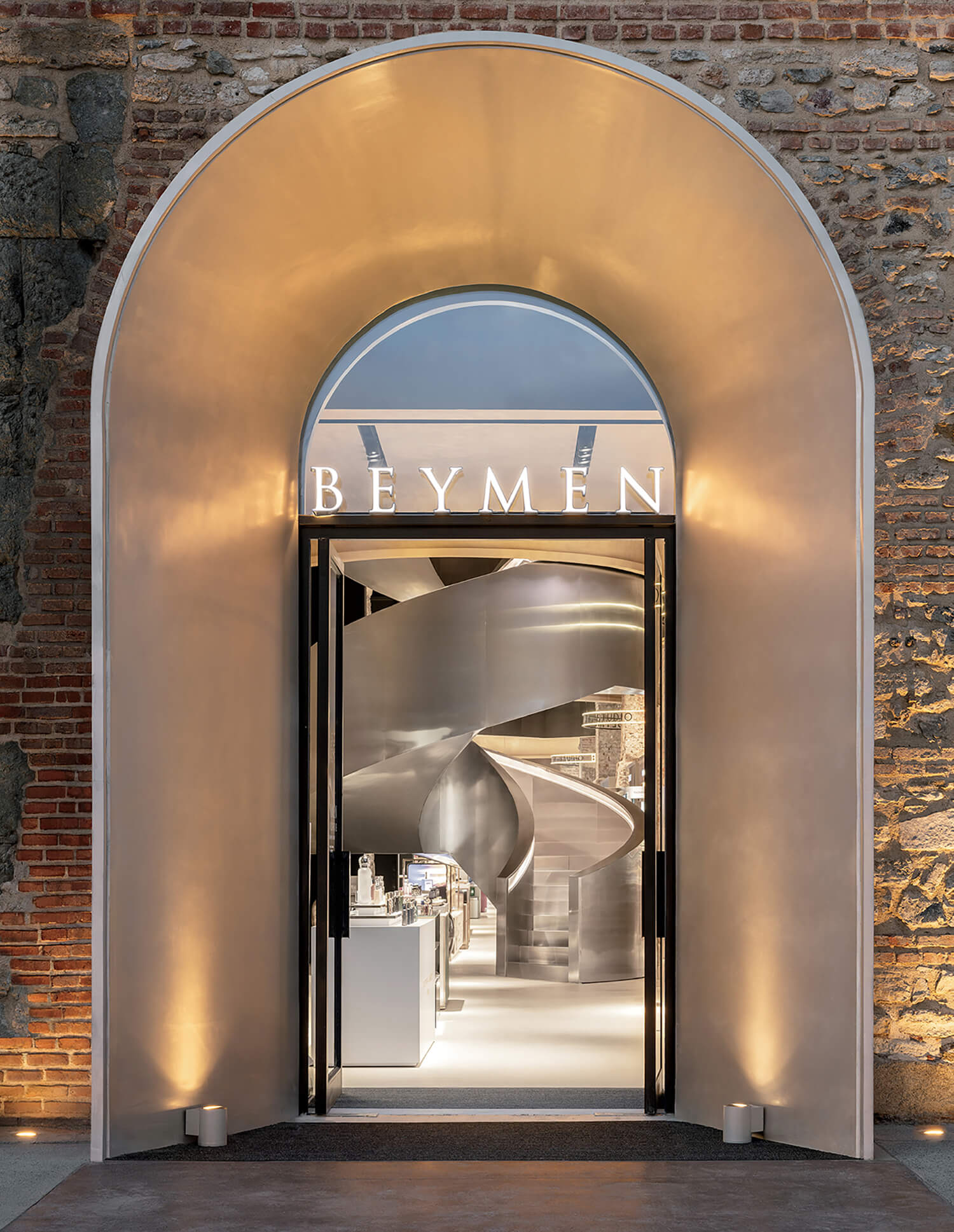 Illuminated entrance to a BEYMEN store set in a brick wall, featuring a large arched doorway and a modern spiral staircase visible inside.