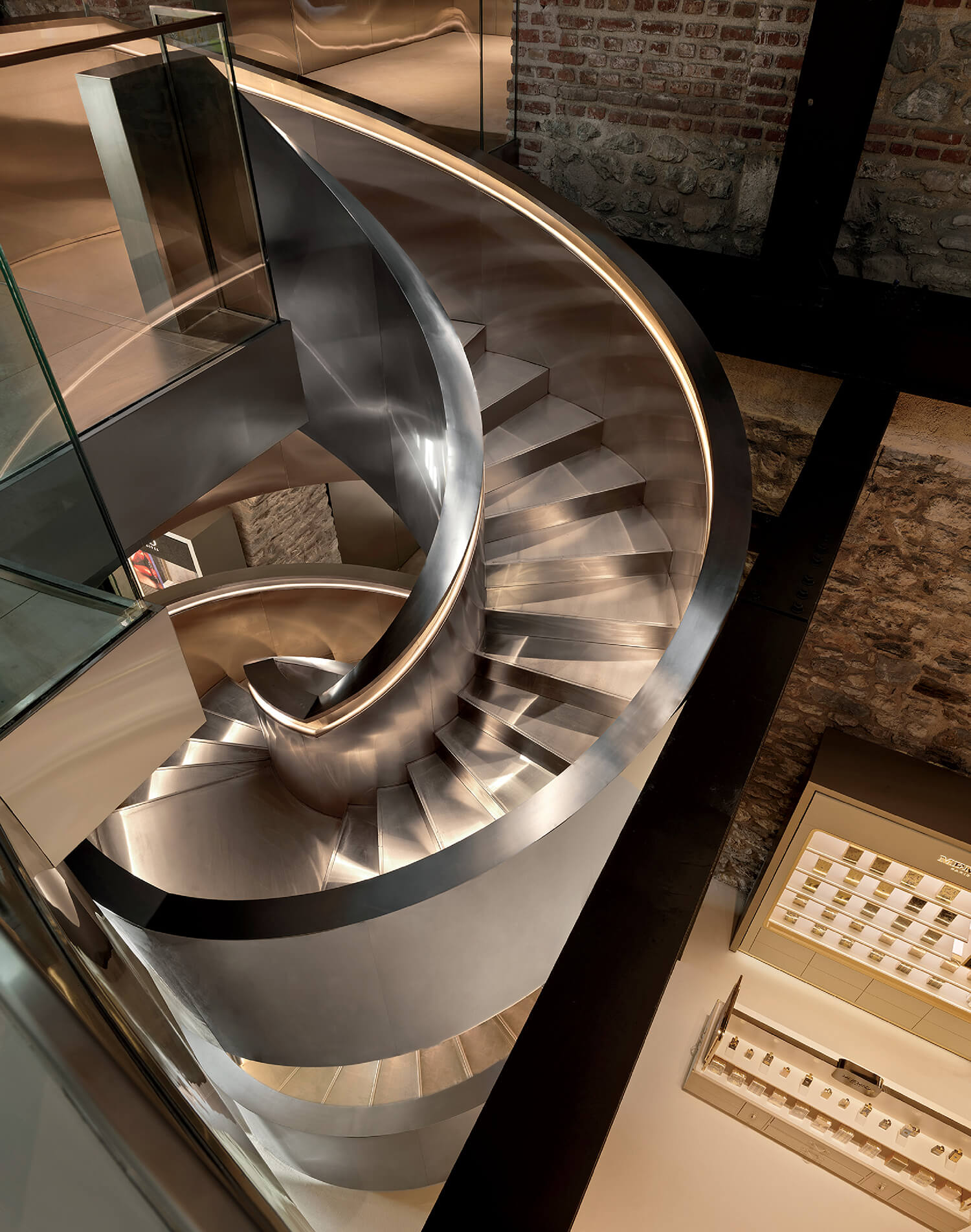 A modern spiral staircase with metallic steps and railing, surrounded by glass and stone walls, viewed from above.