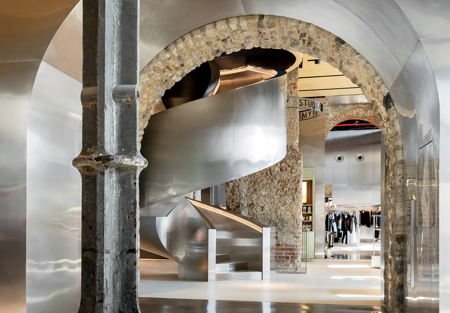 Modern retail space with exposed stone arches, steel columns, and a large metallic spiral staircase; clothing racks visible in the background.