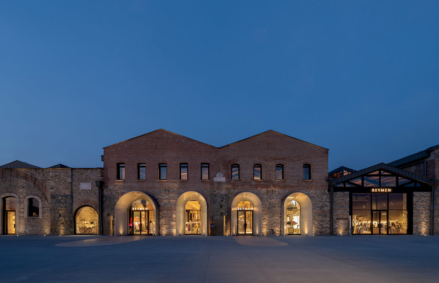 Brick building with large arched entrances and illuminated interior, photographed at dusk under a clear sky.