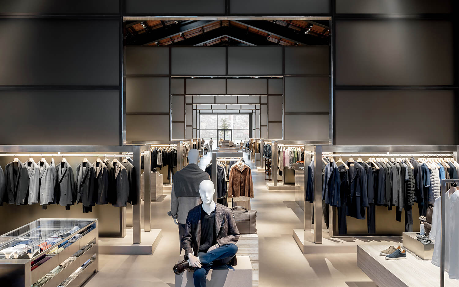 Modern clothing store interior with mannequins, racks of suits, shirts, and jackets, display cases, and shelves with shoes under bright lighting.