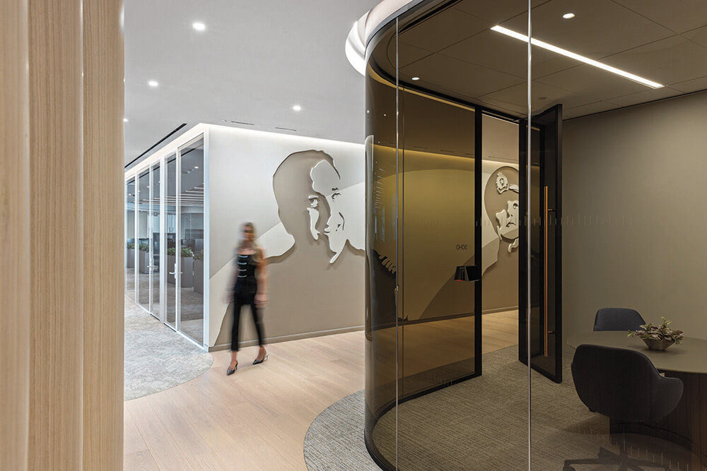 Modern office interior with glass walls, wall art featuring faces, and a person walking through a corridor with wood flooring and contemporary decor.