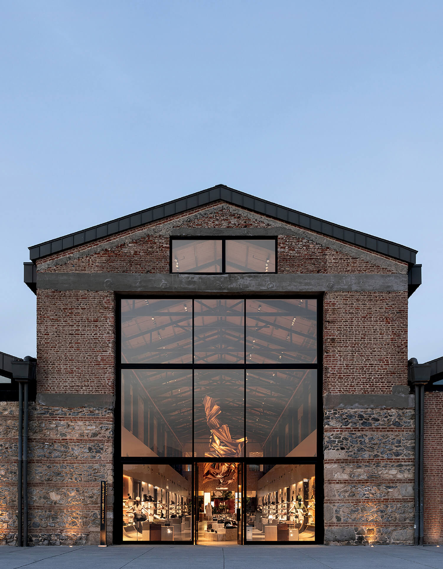 A brick building with large glass entrance doors reveals a modern, well-lit interior with a sculpture and retail displays inside.