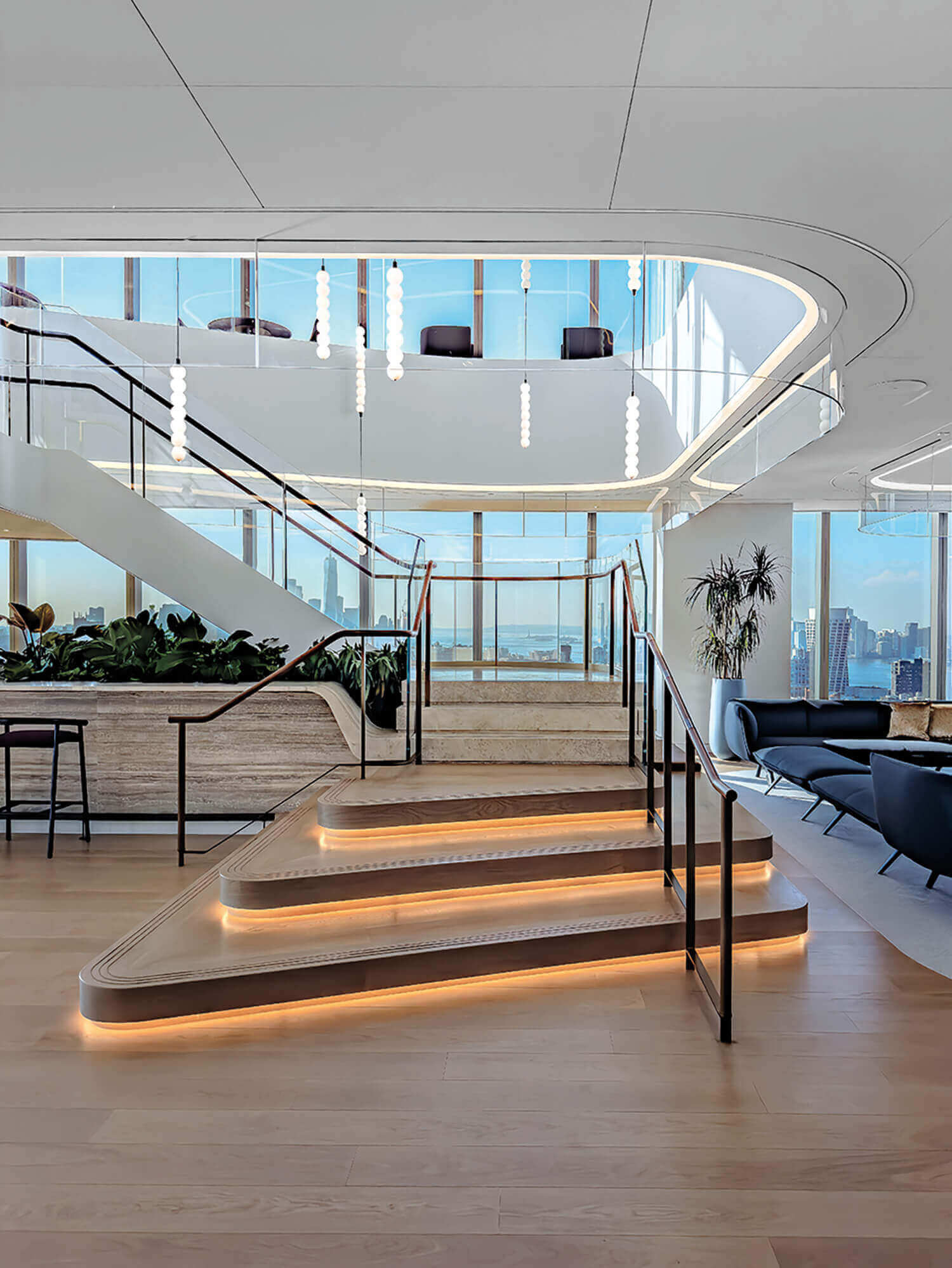Modern office interior with illuminated stairs, glass railings, lounge seating, large windows, and city skyline views.