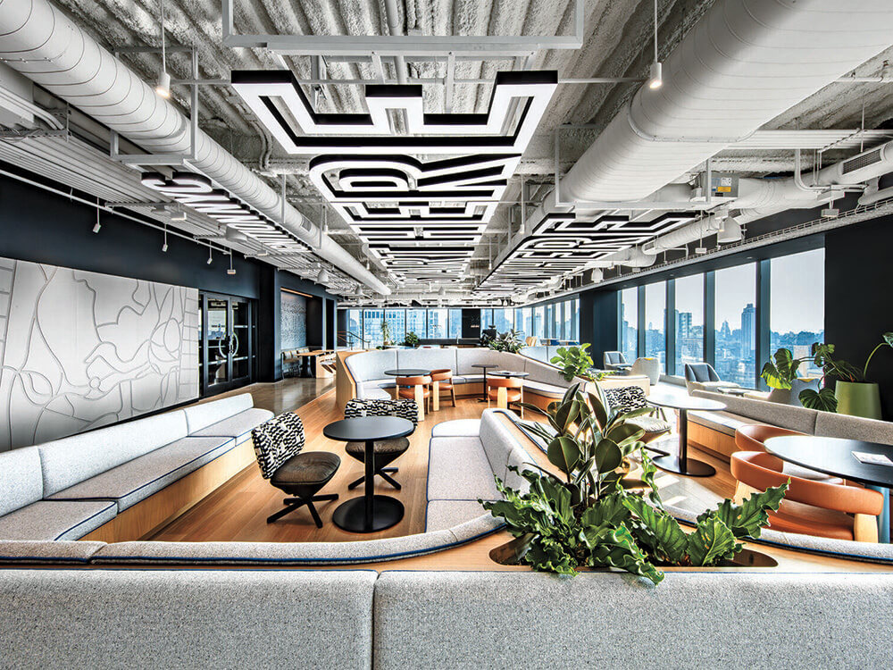 Modern office lounge with curved gray seating, round tables, decorative plants, large windows, city views, and bold geometric patterns on the ceiling and walls.
