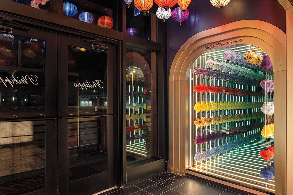 A colorful wall display of umbrellas in a mirrored alcove, next to glass doors and black tile flooring, with lanterns hanging from the ceiling.
